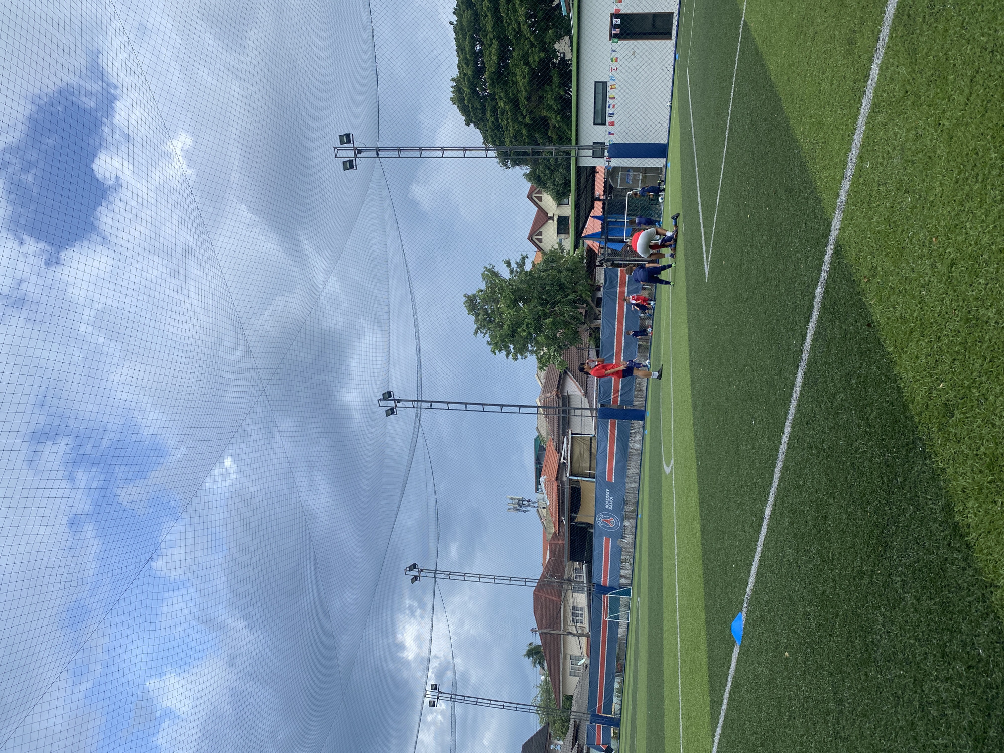 The image depicts an outdoor artificial turf soccer field in a somewhat overcast urban environment. A soccer academy banner is visible along the side of the field, featuring the Paris Saint-Germain logo and the words "ACADEMY SAMUI". Several individuals, appearing to be young players and possibly a coach, are engaged in activities on the field. Some are standing, while others are in dynamic poses, suggesting practice or training. One person is on the ground, receiving attention or assisting another individual who is also on the ground. The field is marked with white lines and blue cones are placed on the turf. Tall light poles with floodlights are positioned around the field, and a safety net stretches overhead. In the background, residential buildings with red-tiled roofs are visible, along with trees and some cellular antennas. The overall scene suggests a sports facility operating in a residential area.