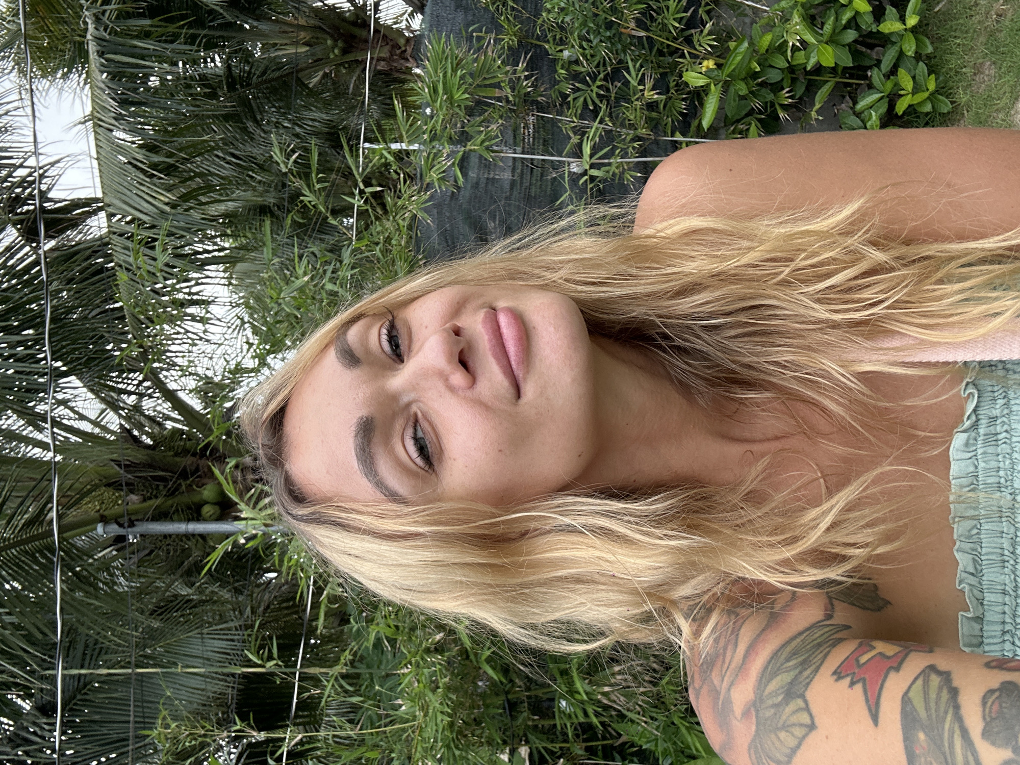 This image is a close-up portrait of a young woman with long, wavy blonde hair, taken outdoors against a backdrop of lush greenery. The photograph is rotated 90 degrees clockwise.
The woman has fair skin, visible freckles, and light-colored eyes. Her eyebrows are dark and well-defined, and her full lips are tinted with a light pink lip gloss. She gazes slightly off-camera with a neutral, calm expression. Her left arm, prominently featured, is covered in extensive, colorful tattoos. She is wearing a light-colored top, possibly light blue or green, featuring ruched detailing across the chest.
The background is rich with tropical foliage, including large palm trees with visible fronds and small green coconuts, alongside dense clusters of what appears to be bamboo or tall grasses. Thin wires or strings are subtly visible crisscrossing through the background vegetation, hinting at a fence or a cultivated area. The bright, diffused lighting suggests it is daytime. The setting, characterized by its abundant tropical plants, aligns with the coastal environment of Mũi Né, Vietnam. The image captures a serene moment, highlighting the woman's features within a vibrant natural landscape.