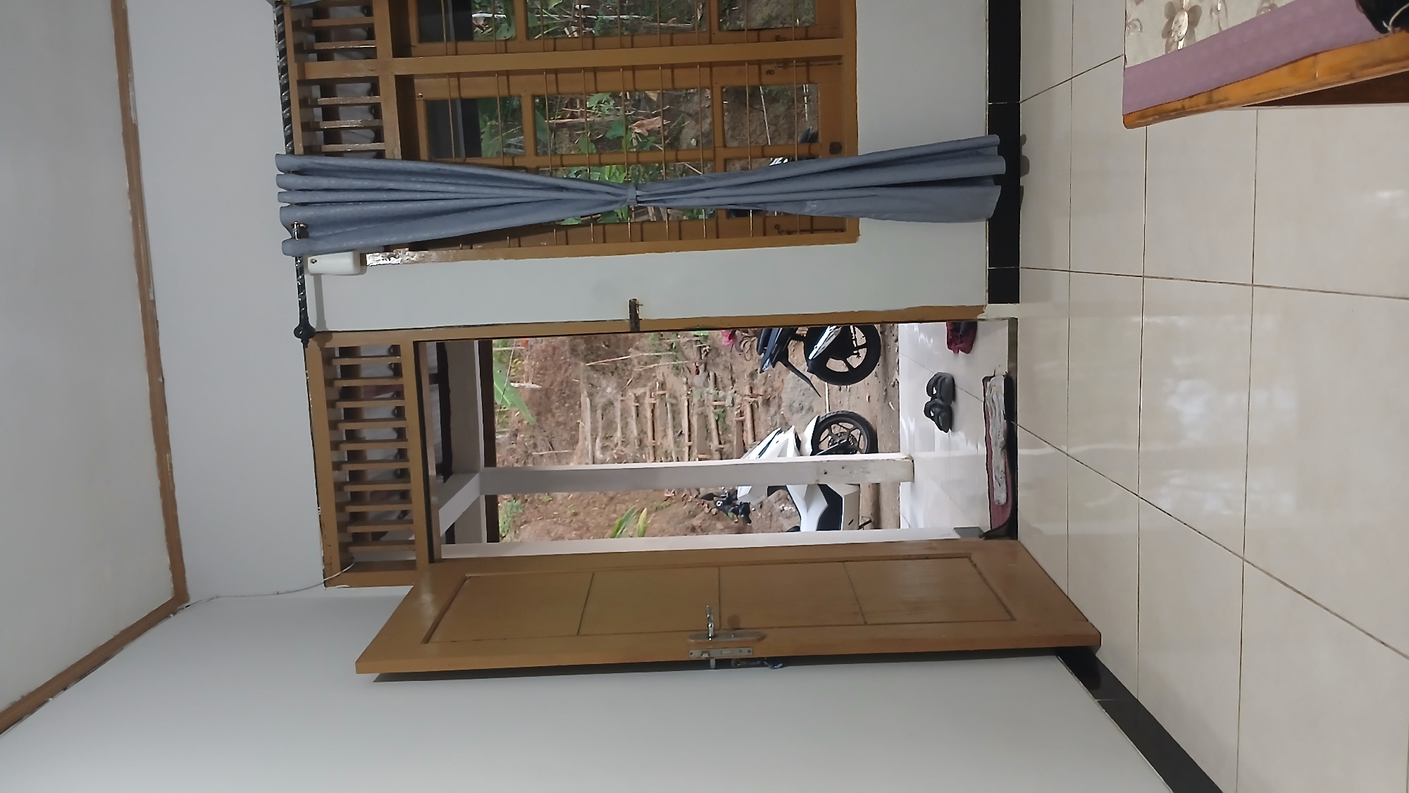 The image shows a view from inside a room with white walls and a tiled floor, looking out through an open doorway towards an outdoor area with stairs leading up a hillside. There is a wooden door ajar on the left, revealing a white motorcycle and a dark-colored motorcycle parked outside. A gray curtain hangs on the right side of the frame, partially obscuring a wooden-framed window. Two pairs of dark-colored sandals are visible on the floor just outside the doorway. The scene appears to be a residential area, possibly in Sampang, Indonesia, given the context.