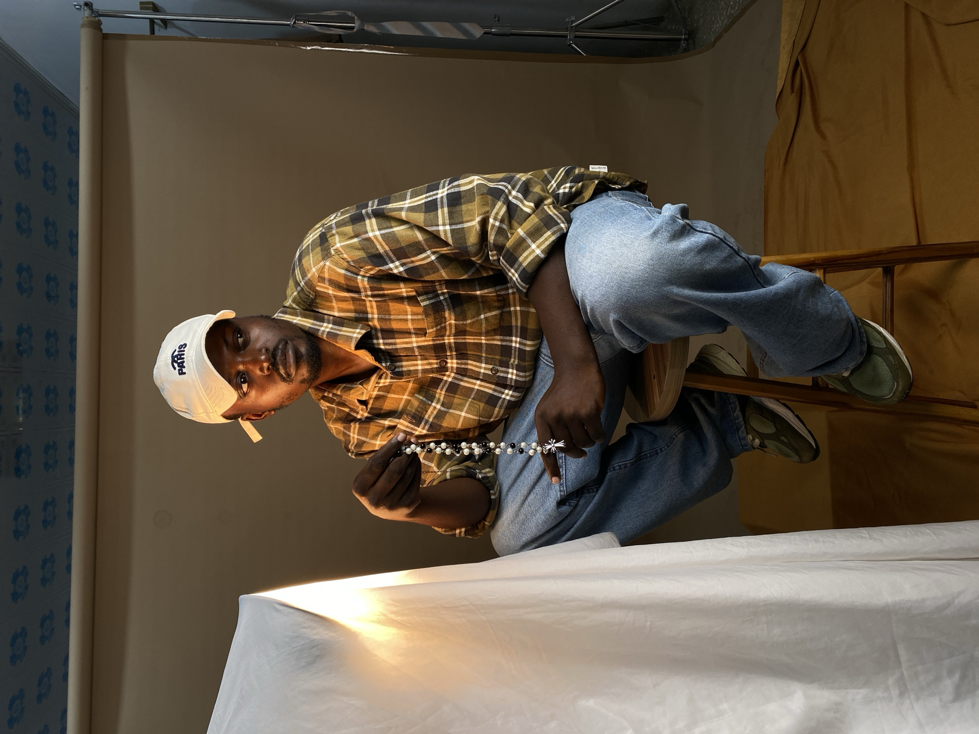 A man wearing a white baseball cap with "PARIS" embroidered on it, a plaid long-sleeved shirt, and blue jeans is sitting on a stool in what appears to be a studio. He is holding a rosary with black and white beads and a silver cross pendant. To his left, a white drape is illuminated by a light source. The background consists of a brown backdrop and a portion of a blue, patterned ceiling. A camera stand is visible on the right side of the image. The setting does not provide any specific clues to identify the location as Gura Top, Nigeria.