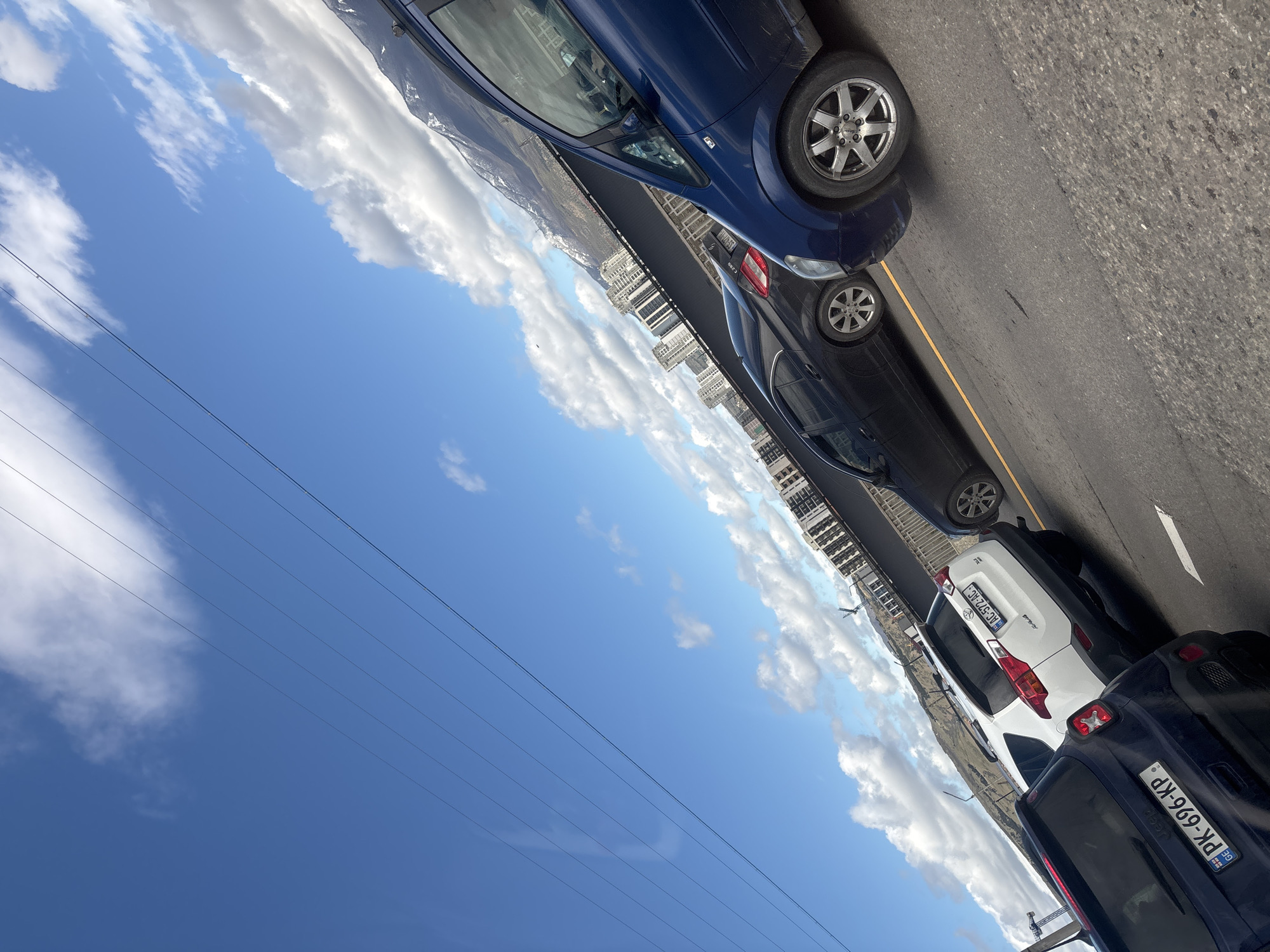 The image shows a street scene in Tbilisi, Georgia. Several cars are parked along the roadside, including a dark blue sedan, a black sedan, a white SUV, and a dark SUV. The dark SUV has a license plate reading "PK-696-KP." The license plate of the white SUV reads "BAC-572-AC." Buildings and a mountain range are visible in the background, suggesting the location is in an urban area with nearby natural features. The sky is blue with scattered clouds. Power lines are visible stretching across the sky. The scene appears to be captured during daylight hours.
