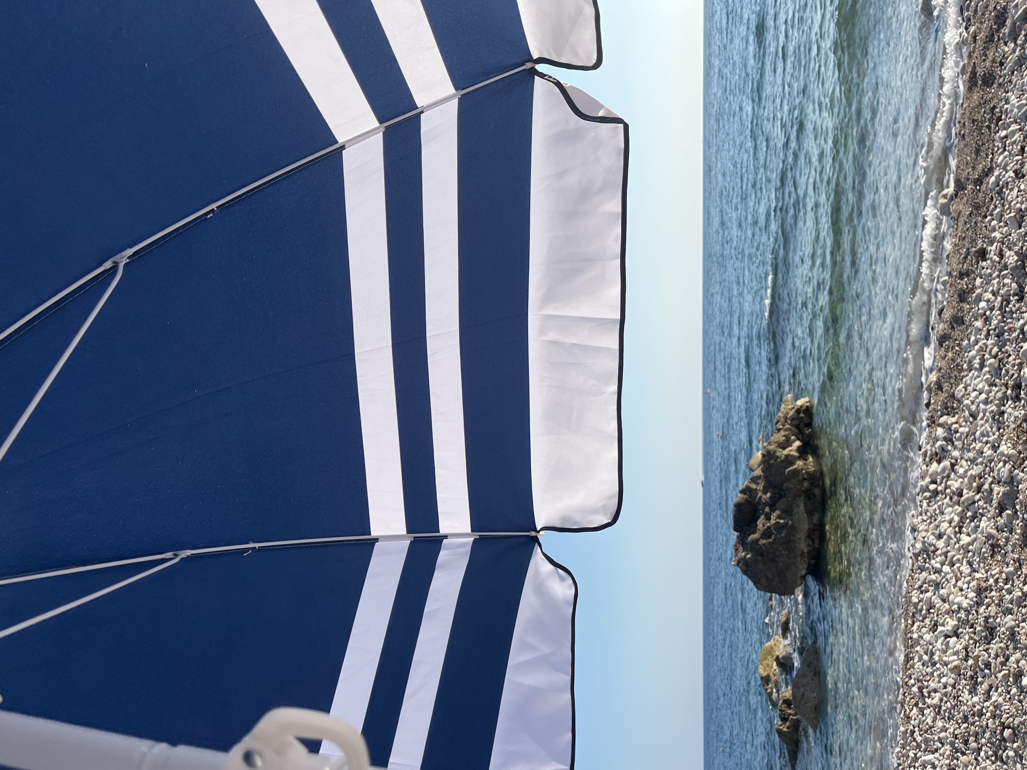 An upward angled shot captures the underside of a blue and white striped beach umbrella casting a shadow over a clear, pebble-strewn beach and the Adriatic Sea. The water is calm with gentle waves lapping the shore, and a few large rocks are visible near the waterline. In the distance, the horizon is clear, meeting a pale blue sky. The scene is set in Bar, Montenegro, on a sunny day.