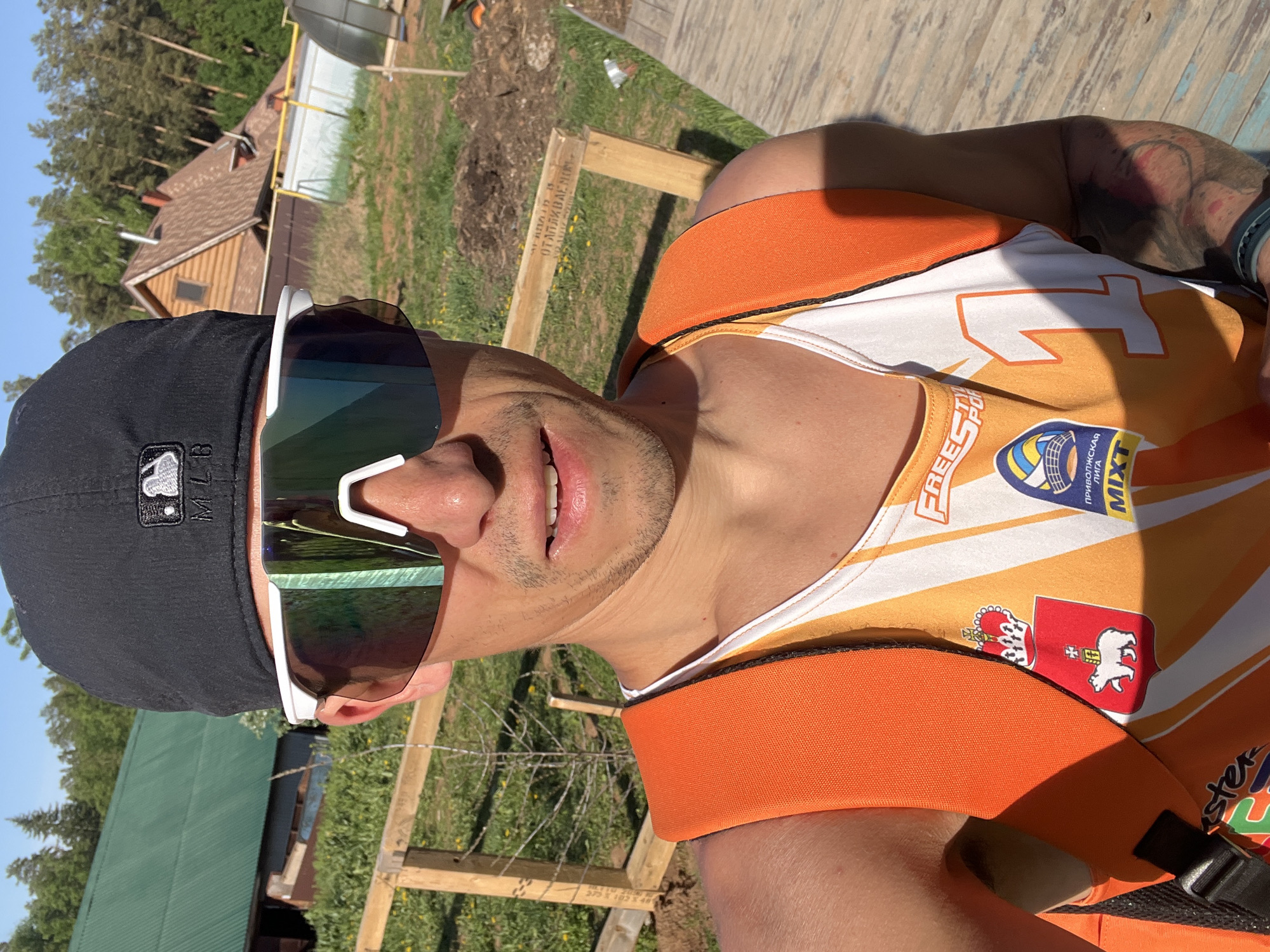 A man wearing a black baseball cap, white and green sunglasses, and an orange and white sleeveless jersey with various logos is standing outdoors. The jersey has a red and white emblem with a bear and a crown on the lower left. Behind the man, there's a grassy area with wooden structures, including a fence. A wooden cabin is visible in the background. The location is indicated to be in Chaikovsky, Russia based on the logos present on his jersey. The man has a light stubble.