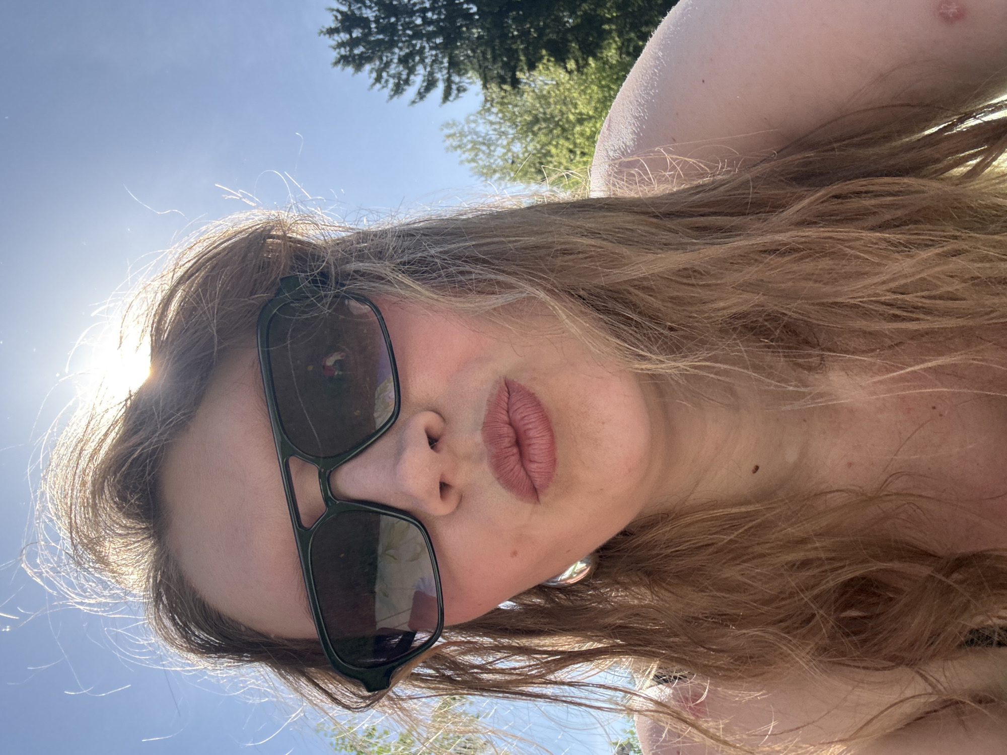 A woman is photographed from a low angle, looking down at the camera. She wears sunglasses and has long, light brown hair. The sky is visible in the background, along with some trees. The location is not discernible as being in Izhevsk, Russia.
