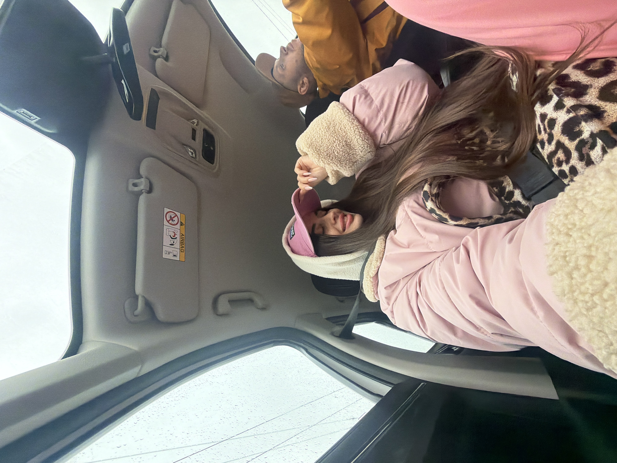 The image depicts the interior of a car, possibly a sedan, with three people inside. The person in the foreground, a woman, is wearing a pink jacket with a sherpa-lined collar and a pink baseball cap. She has long, brown hair. Adjacent to her, a pillow with an animal print (leopard or cheetah) is visible. Behind the woman, a man is partially visible, wearing a brown jacket. The car interior features light-colored upholstery. The car's windshield is visible with raindrops on it, suggesting wet weather conditions. The environment outside the car is not clear. The location, Saint Petersburg, Russia, is not definitively discernible from the visual information provided.