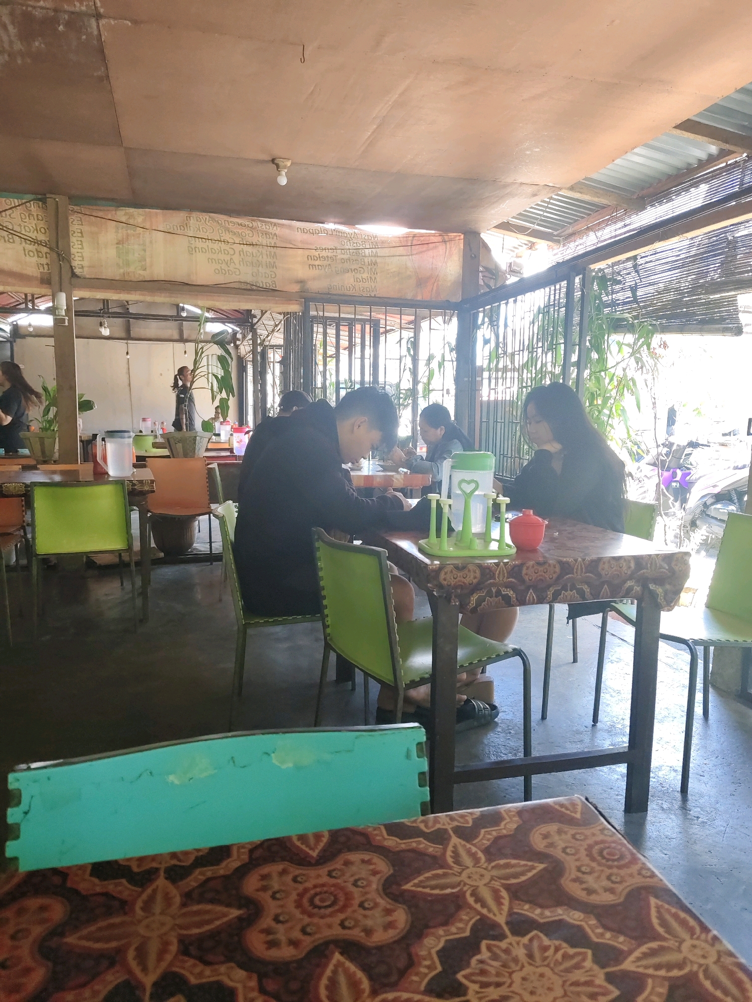 The image depicts an outdoor dining area, likely a warung, in Tomohon, Indonesia. In the foreground, a patterned tablecloth is visible on a table. Behind this, four individuals are seated at a table. Two of them, a man and a woman, are the most clearly visible. The man, on the left, is wearing a dark jacket and is looking down. The woman, on the right, has long dark hair and is also looking down, appearing to be engaged with her phone. A green pitcher and a red bowl are on the table. The environment is rustic, with a corrugated metal roof overhead and metal fencing. Green chairs are scattered throughout the seating area. Sunlight streams in, casting shadows. In the background, more people are present, and plants are visible. A purple motorbike is parked to the right. A banner with text, likely a menu or advertisement, is hanging on the wall. The overall impression is of a casual, open-air eatery.