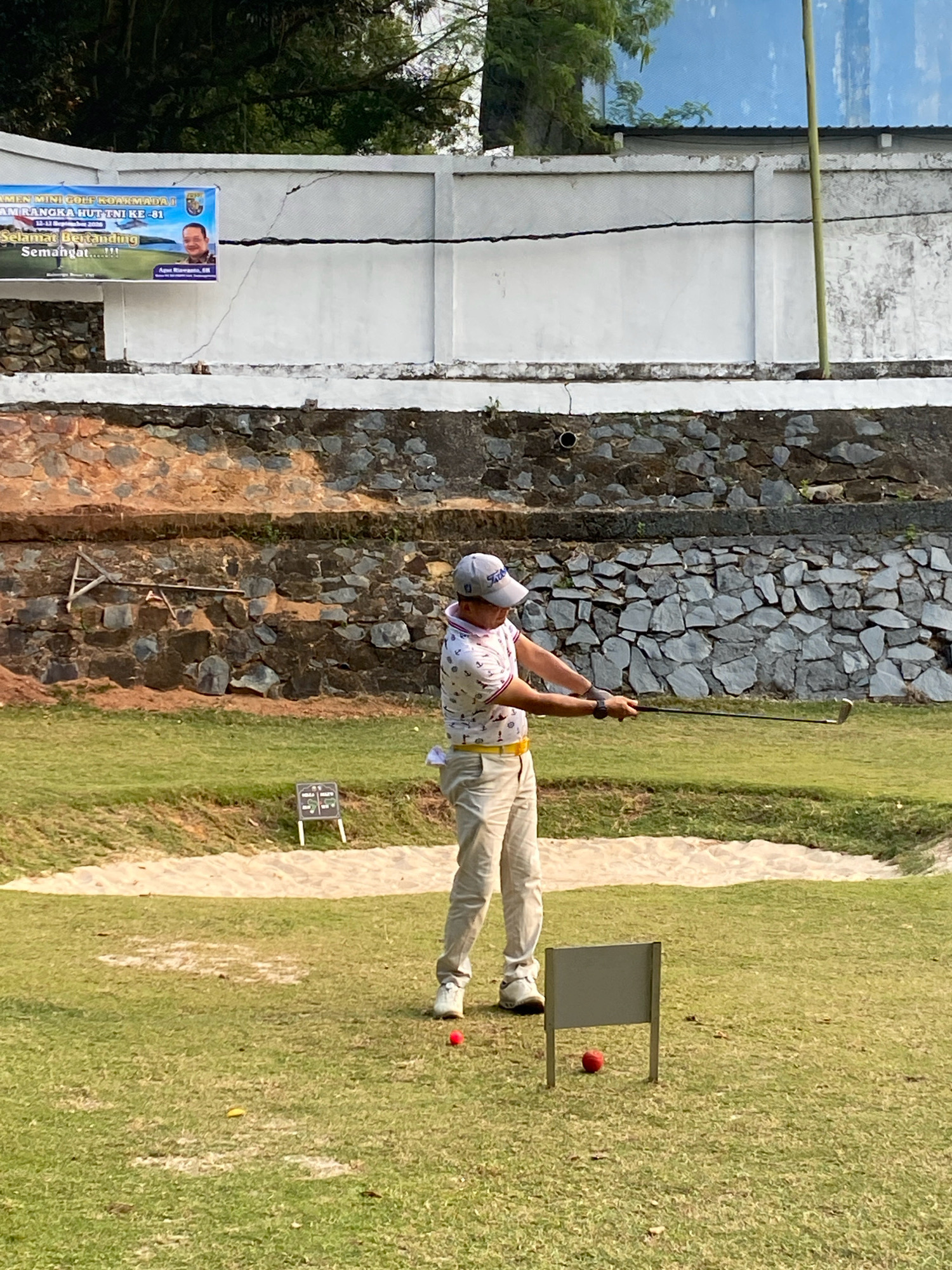A male golfer, wearing a white polo shirt with a pattern of palm trees, light beige pants, and a grey baseball cap, is in the process of swinging a golf club on a grassy golf course. He is positioned within a sand bunker, with a sand trap behind him and the fairway in front. The golfer's stance suggests he is about to hit a golf ball, which is not clearly visible in relation to his club.
In the foreground, two small grey signs are planted in the grass, one with a red golf ball in front of it. Another red golf ball is visible on the grass to the left of the golfer. The golf course appears to be situated in a hilly area, with a stone wall constructed on a slope behind the golfer. Above the stone wall, a white concrete wall with a banner attached is visible. The banner, partially obscured by shadows, appears to advertise a mini golf event related to the Indonesian Navy's 81st anniversary, with text in Indonesian and a picture of a man. The overall environment suggests a sunny day, with some trees visible in the background. The location is identified as the Rinjani Golf Course in Tanjung Pinang, Indonesia.