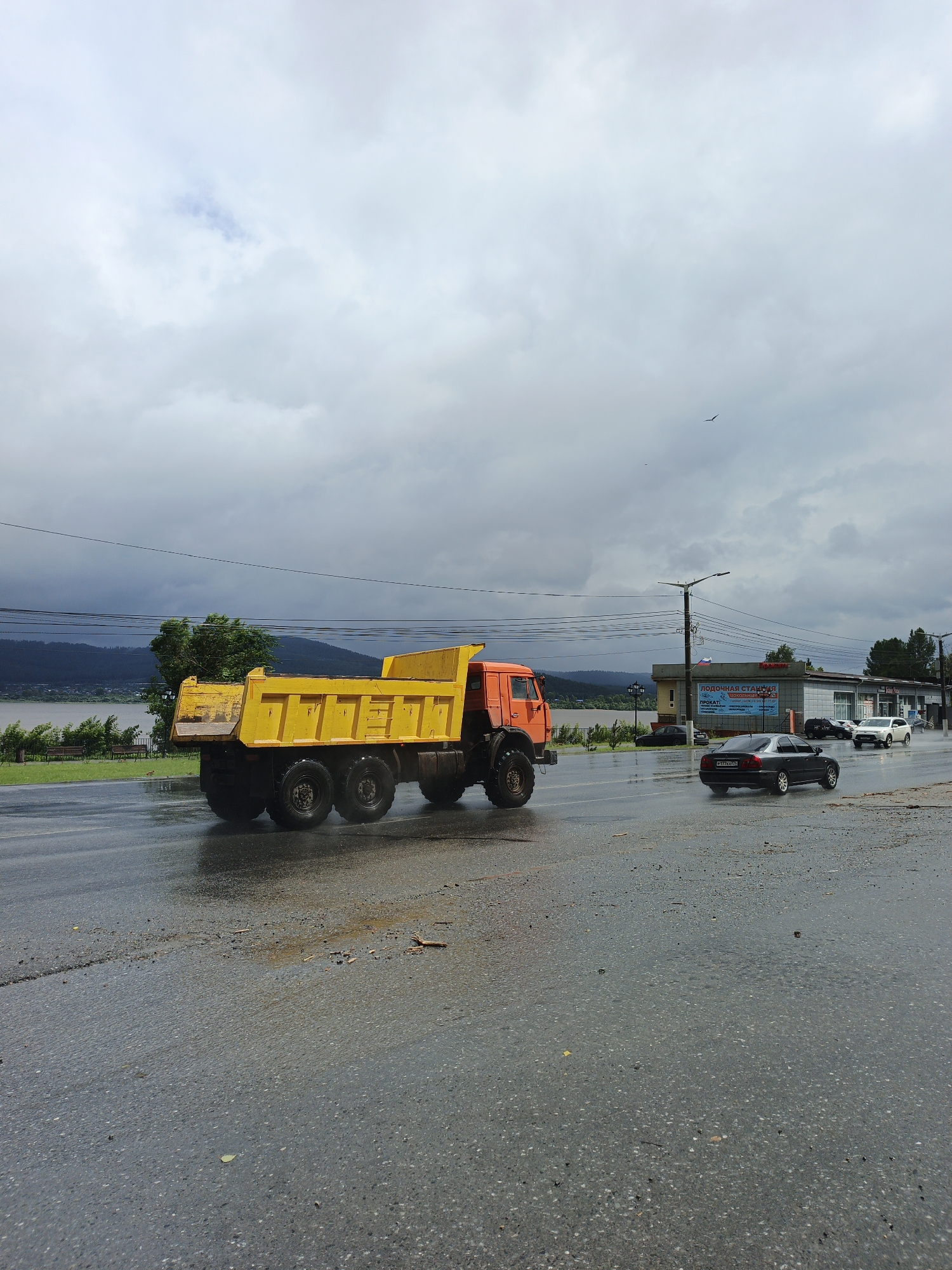 This image depicts a wet asphalt road with a large orange and yellow dump truck on the left, its rear wheels splashing water. Several cars, including a black sedan and a white SUV, are driving on the road to the right. In the background, there is a low-rise building with signage that appears to be in Russian, suggesting a commercial establishment. Beyond the building, a body of water and tree-covered hills are visible under a cloudy sky. The presence of the building and the signage, combined with the geographical features, suggests this scene is likely located in the city of Zlatoust, Russia, possibly near a waterfront area. The overall impression is of a day with inclement weather, likely rain, given the wet road surface and splashing water. There are no people clearly visible in the image.