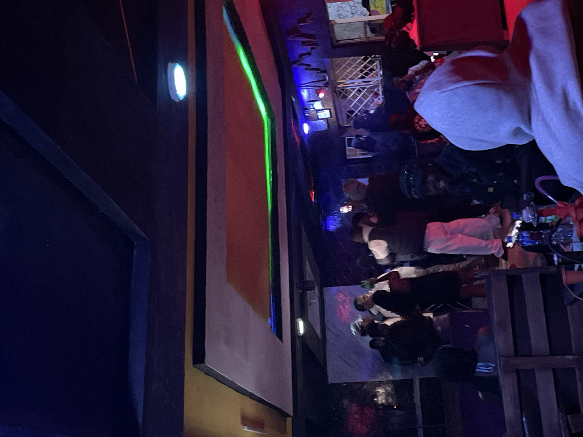 The image depicts an indoor gathering at a nightclub or lounge in Durumi, Nigeria. The environment is dimly lit, with vibrant neon lighting contributing to the atmosphere. The ceiling features multi-level designs with yellow and red accents, and a prominent green neon strip runs along one of the upper levels.
In the foreground, a person wearing a light grey hooded sweatshirt is visible from behind, facing away from the camera. To the left, a wooden bench or seating area is present. Further into the scene, several individuals are gathered. Some appear to be standing and interacting, possibly dancing or socializing. One individual in the center is wearing a white top and light-colored pants. Another person to the left is dressed in dark clothing, and another in the middle ground is wearing a dark jacket and a cap.
In the background, there is a wall with decorative elements and some framed pictures. A projection screen or a wall display with colored lights is also visible. The overall impression is that of a lively social event taking place in a modern entertainment venue.
