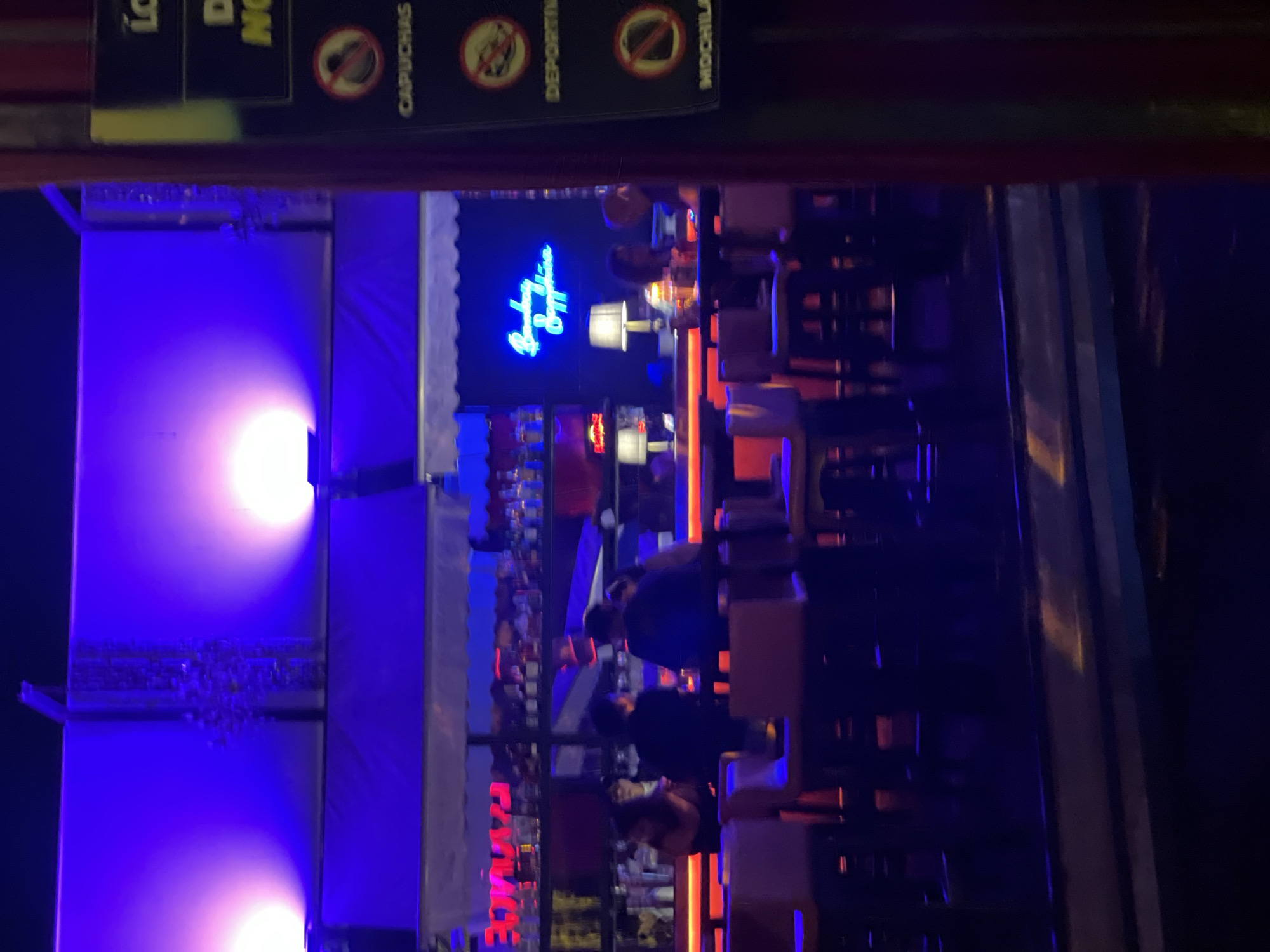 The image shows an outdoor bar or restaurant in Arica, Chile, at night. There are customers sitting at the bar, and staff serving them. Neon lights illuminate the bar and the sign above it, with a menu visible at the top. The lighting is blue and orange. There are people at the bar and sitting at tables.