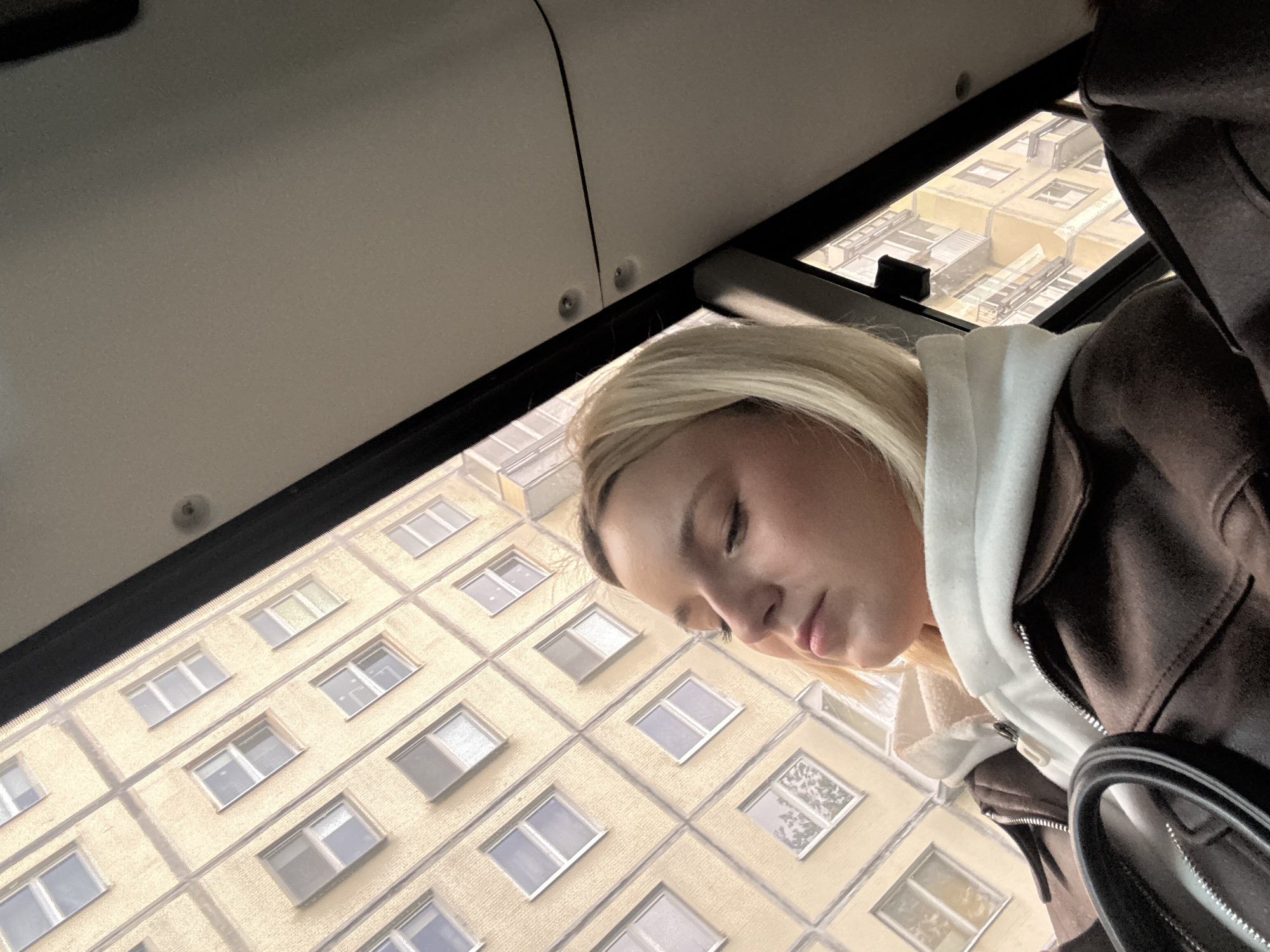 A young woman with blonde hair and closed eyes is positioned in the right foreground of the image, seemingly asleep. She is wearing a white hooded sweatshirt under a brown jacket, with part of a handbag visible on the right. The background consists of an apartment building, likely viewed from inside a vehicle. The building is light yellow, with multiple rows of windows, indicating a multi-story structure. The perspective suggests the photograph was taken from inside a bus or car. The upper left portion of the image shows the interior of the vehicle with visible ceiling and a black frame structure. Given the information provided, the location is potentially in Sinyavino, Russia.