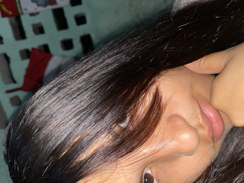 A close-up selfie focuses on a woman's face, tilted towards the camera. Her straight, dark hair partially obscures her face, with a few strands falling over her eye. The visible eye is brown, and her skin appears smooth. Her mouth is slightly open, and one hand is touching her face near her lip. The background features a light blue wall with square cutouts, and a glimpse of red and white fabric is visible in the upper left corner.