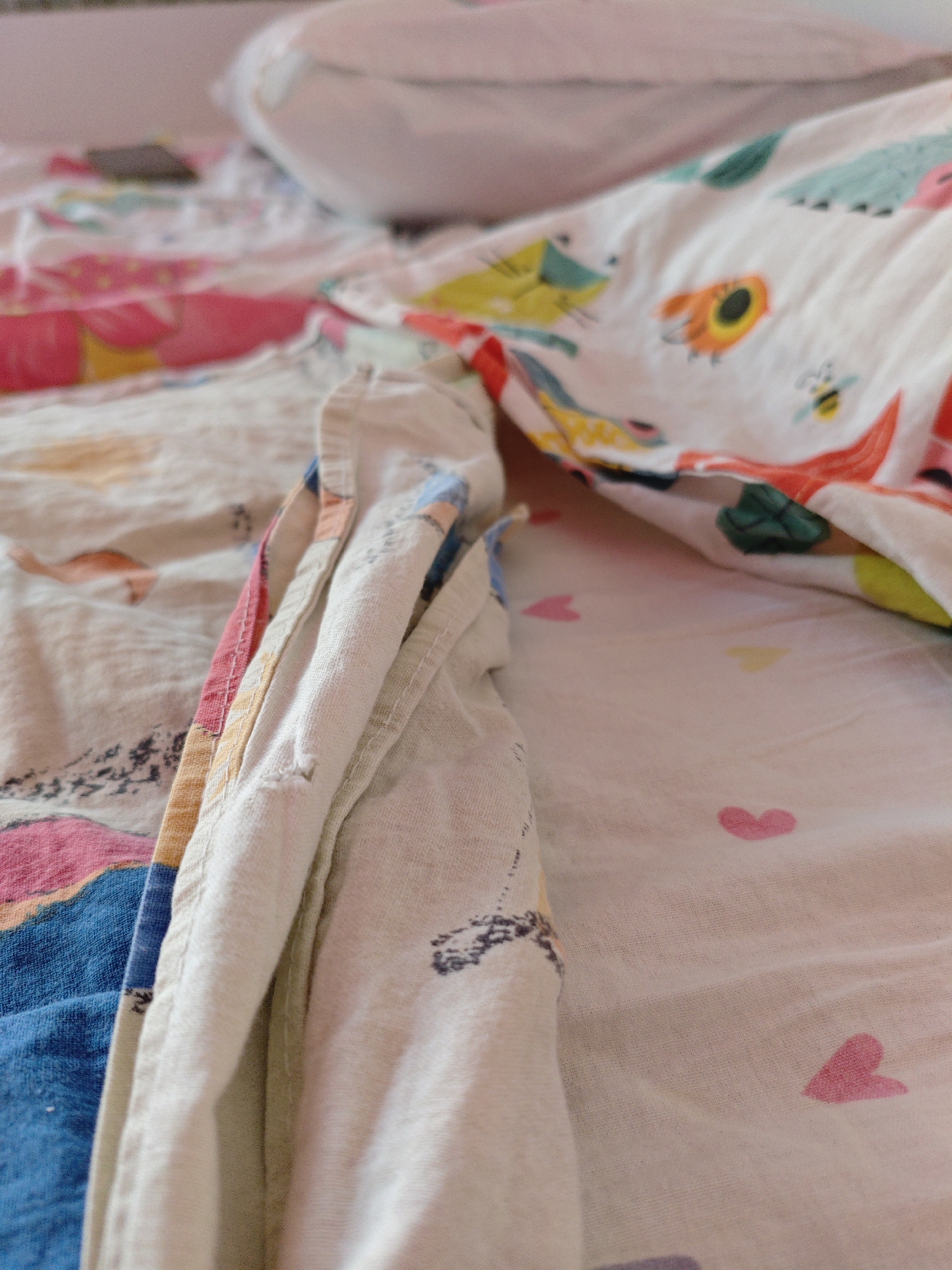 The image shows a close-up view of a bed with colorful, patterned bedding. The bedding features cartoonish animals like cats and bees, as well as small pink hearts scattered across a white background. A crumpled duvet is prominent, with a section folded over, revealing a seam with pink, yellow, and blue trim. The scene appears to be indoors, likely in a bedroom, based on the presence of a bed and pillow. The lighting suggests natural light, possibly from a window. There are no people visible in the image. The setting does not provide any specific geographical clues to pinpoint its location within Трудовское сельское поселение, Ukraine.