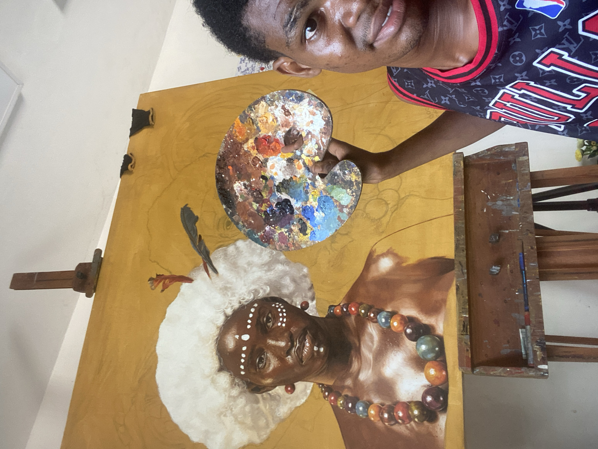 A young, dark-skinned artist is shown from the chest up, holding a paint palette in front of a large, unfinished painting on an easel. The artist, who has short curly hair and a slight smile, is looking towards the camera. He is wearing a black sleeveless top with red trim, patterned with Louis Vuitton monograms and featuring an NBA Chicago Bulls logo and text.
The painting on the wooden easel depicts a highly detailed portrait against a warm, yellow-ochre background. The subject, likely a woman, has dark skin, an elaborate fluffy white headdress or wig with dark feathers, and white dots painted in patterns on her forehead, cheeks, and chin. She wears a vibrant, multi-colored beaded necklace and small red earrings. The subject in the painting has a calm, slightly smiling expression.
The kidney-shaped paint palette held by the artist is covered with various colors of paint, indicating recent use. To the right of the painting, part of the wooden easel is visible, including a small shelf that holds a pen or brush, with the word "ALLSTON" faintly visible on it. The background of the scene appears to be an indoor space with a plain light-colored wall and a yellowish floor. The overall impression is that of an artist presenting his work in a studio setting in Ado, Nigeria.
