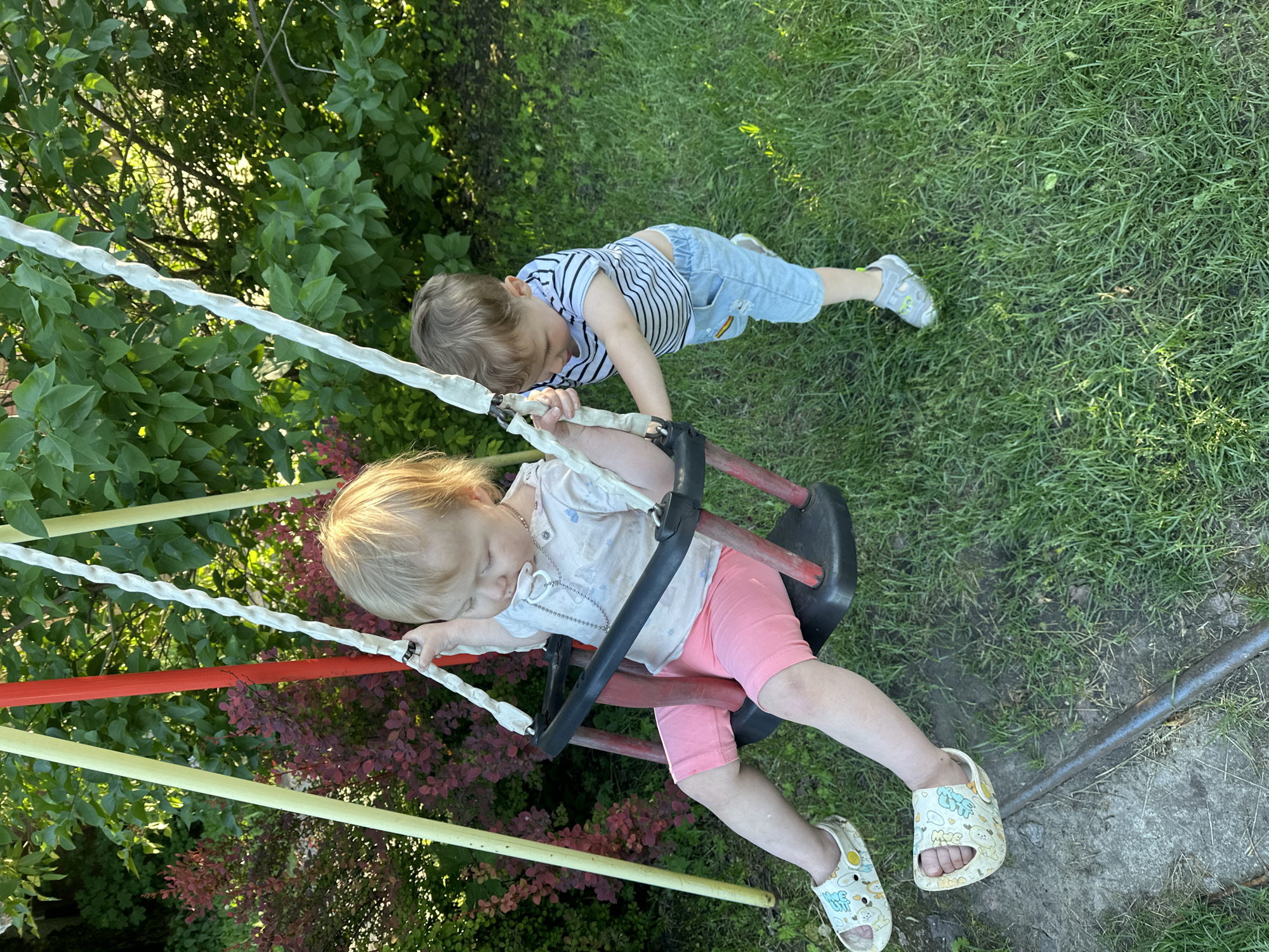 This image depicts two young children interacting with a swing set in an outdoor setting in Nizhny Novgorod, Russia, during daylight hours with bright sun creating distinct shadows. A fair-haired child, appearing to be a girl, is seated in a black plastic swing, suspended by white ropes from a swing set comprising yellow/cream and red poles. She wears a light-colored t-shirt, pink shorts, and light-colored sandals adorned with cartoon characters, and has a pacifier on a chain around her neck. Her gaze is directed downwards. Adjacent to the swing, a brown-haired child, appearing to be a boy, stands and is actively pushing the swing. He wears a horizontal striped t-shirt, light blue denim shorts with a rainbow patch on the left thigh, and grey-green closed-toe sandals/shoes. The ground is primarily covered in green grass, with some areas of exposed dirt and a partially visible metal bar or pipe running across the lower foreground. In the background, dense green foliage from trees and bushes is visible, along with a prominent bush featuring deep red/burgundy leaves.