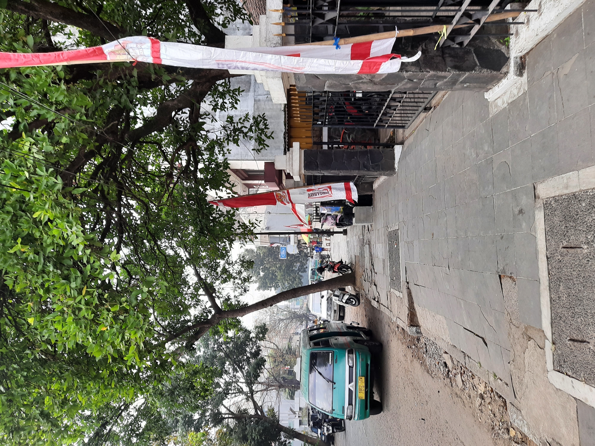The image depicts a street scene in Bandung City, Indonesia, characterized by a narrow sidewalk and a road with parked vehicles. A green minivan is prominently parked on the left side of the road. The sidewalk is made of grey paving stones, some of which are cracked and uneven, indicating wear and tear. A large tree with dense green foliage extends over the sidewalk and road, casting shade.
Flags with red and white stripes are displayed, one of which is attached to a bamboo pole near a black metal fence. The fence encloses a property with a building visible behind it. A few motorcycles are parked along the sidewalk. In the background, other vehicles and buildings can be seen, suggesting a residential or commercial area. The overall atmosphere is that of a typical urban street in Indonesia.
