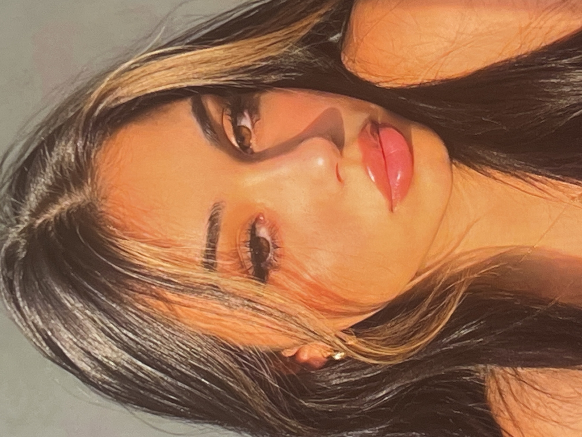 A close-up portrait of a young woman is captured, bathed in warm, golden-hour lighting. Her dark hair, streaked with lighter brown highlights, frames her face, with strands falling forward to partially obscure her left eye and shoulder. She wears a small, gold hoop earring in her visible left ear. Her makeup is prominent, featuring dramatic winged eyeliner, full lashes, and a glossy pink lipstick that accentuates her full lips. The warm light casts a strong shadow across the left side of her face, highlighting her cheekbone and nose. The background is a muted, neutral gray, providing no specific environmental context.