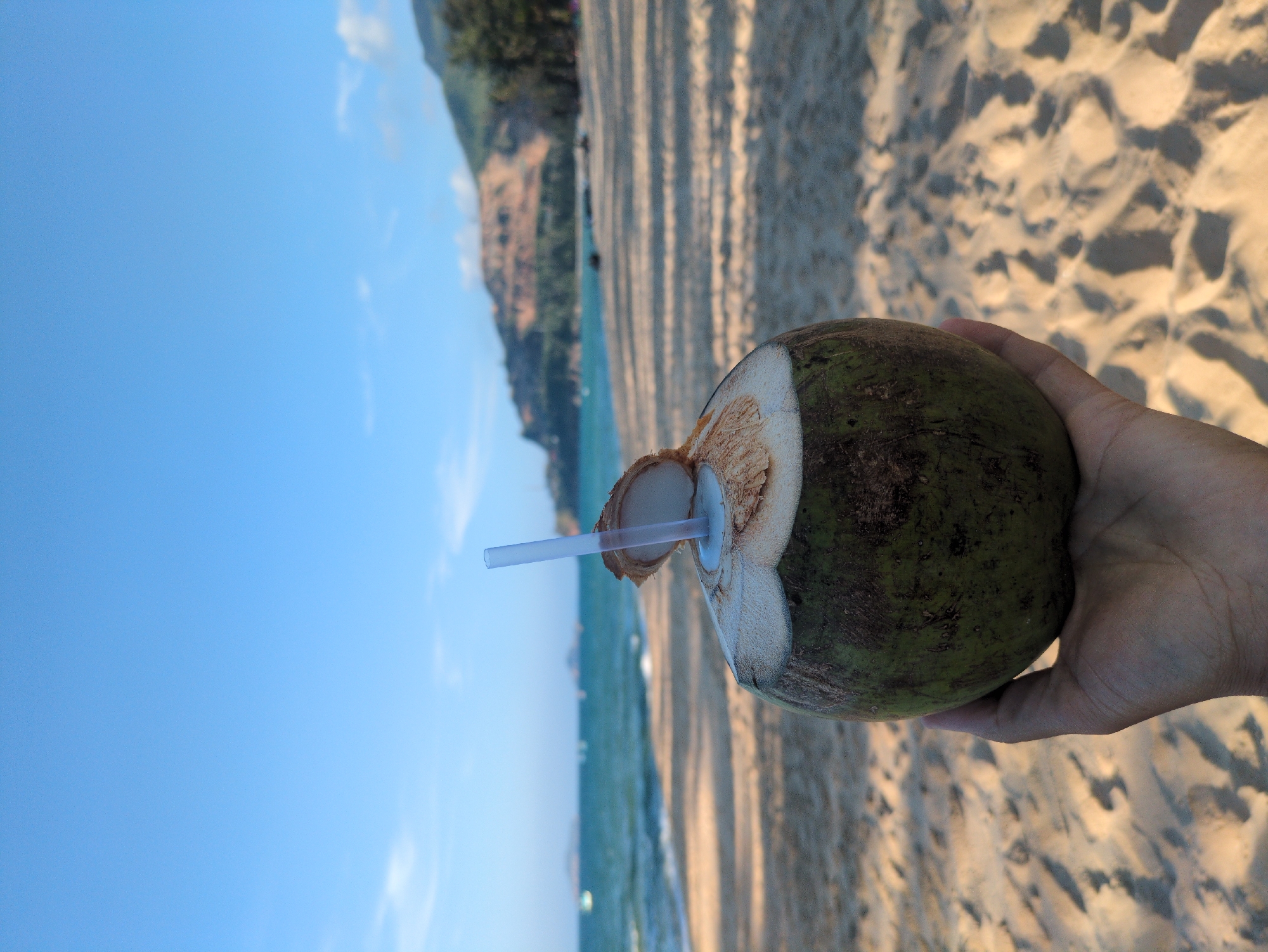 A coconut with a straw sits in a hand against the backdrop of a beach and the sea, taken in South Quy Nhon Ward, Vietnam. The foreground shows a hand holding a green coconut with a white straw inserted. The beach is visible beneath the coconut, revealing fine sand with shadows. In the distance, a turquoise sea meets the horizon under a clear blue sky with faint clouds. A strip of land with vegetation sits on the horizon line.