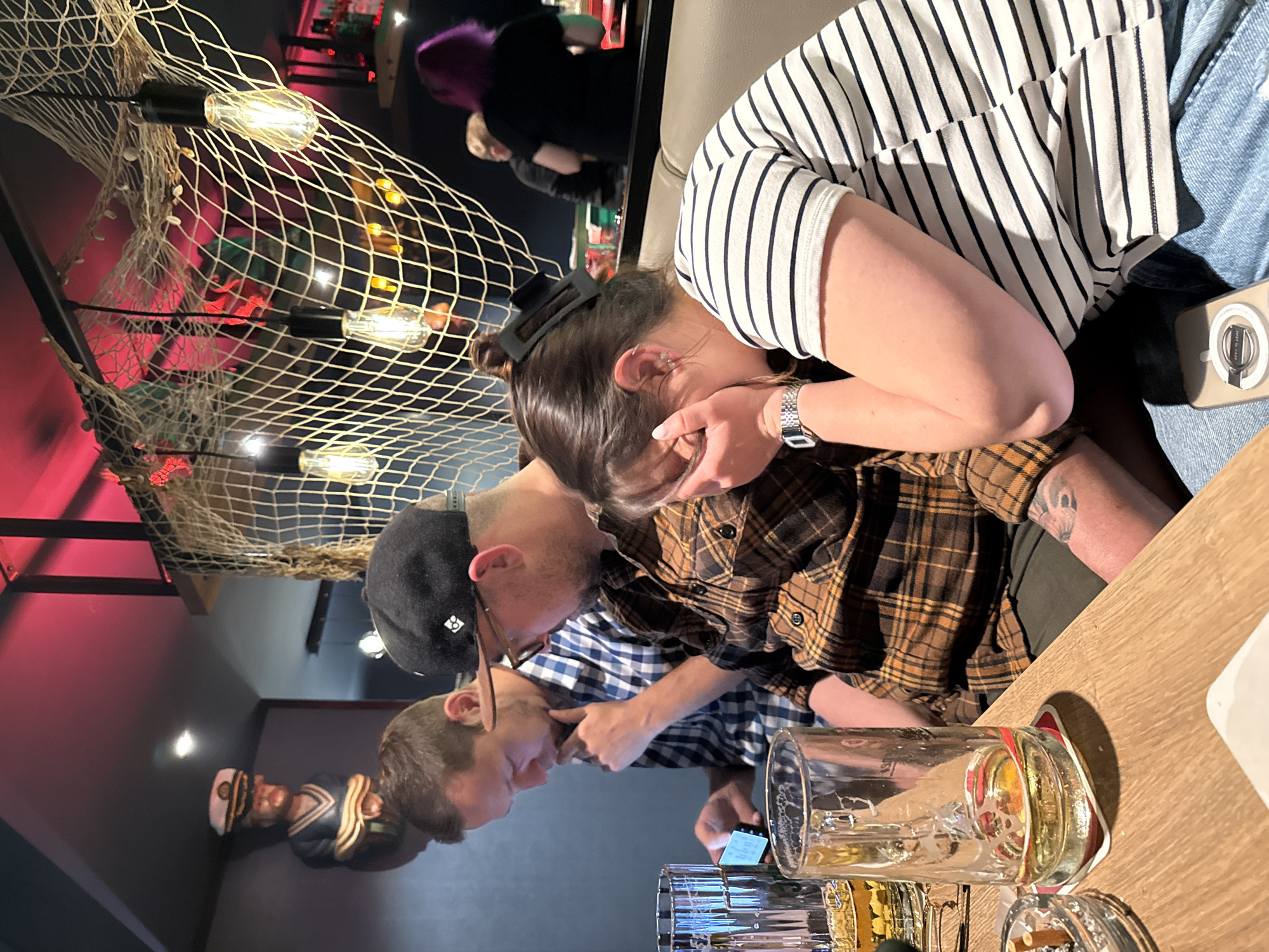The image captures an indoor scene at a maritime-themed bar or restaurant, potentially in the city of Langeoog, Germany, as suggested by the context. Three individuals are the primary subjects. In the foreground, a woman with dark hair, wearing a black and white striped shirt and blue jeans, is seated and covering her face with her hands, her body angled away from the camera. To her left, a man in a brown and black plaid shirt, wearing glasses and a black baseball cap, is seated and appears to be looking at something off-camera. Another man, wearing a blue and white checkered shirt, is seated to the left of the man in the plaid shirt, and is also looking down at a mobile phone he is holding in his hand.
On the wooden table in front of the seated individuals are several items: an ashtray with cigarette butts, a glass of beer, and an empty beer glass. In the background, a decorative net with seashells and lightbulbs hangs from the ceiling, contributing to the maritime theme. A carved wooden figure of a sailor in a captain's hat is visible on a shelf or wall to the left. Further in the background, other patrons and bar elements are partially obscured. The lighting is dim, with red and pink hues from overhead lights illuminating parts of the scene.