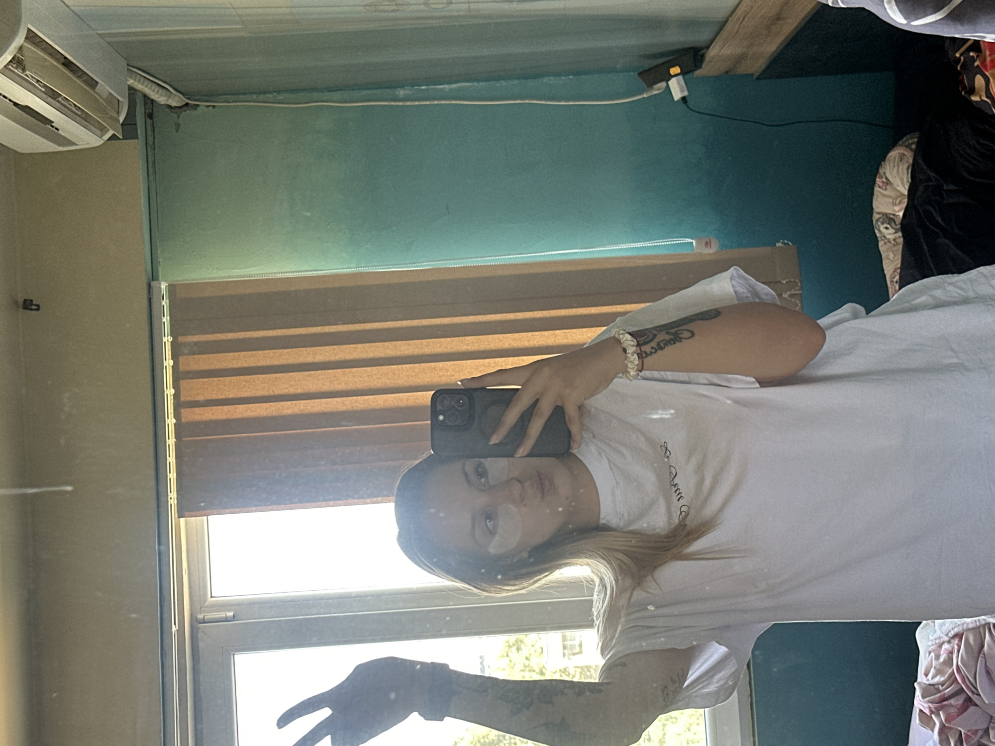 A young woman with blonde hair and under-eye patches is taking a selfie in a mirror. She is wearing a white t-shirt and has her left arm raised with two fingers extended in a peace sign. Her lips are puckered. The background shows a window with vertical blinds, an air conditioning unit, and a teal-colored wall. The image appears to be taken indoors, possibly in a bedroom or living space. The location of Plovdiv, Bulgaria is not evident from the image.