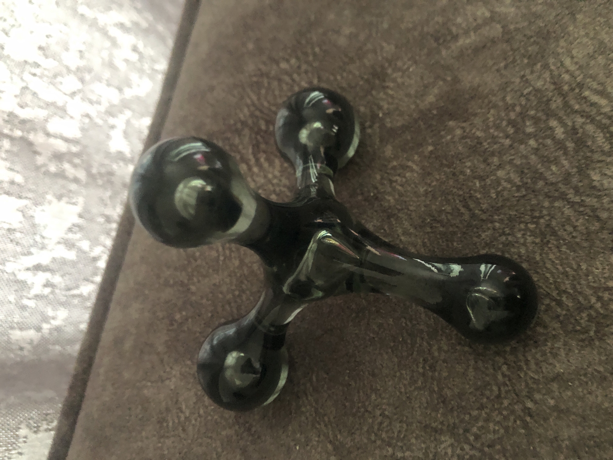 A dark, translucent, cross-shaped object made of glass or resin lies on a brown textured surface, possibly upholstery. The object has a central stem with three rounded protrusions, one at the top, one to the left, and one to the right. The surface it rests on appears to be a couch or chair, with a blurred, light-colored fabric in the background. The lighting casts reflections on the object, highlighting its smooth, polished surface. The location of Путёвка, Russia, is not depicted in this image.