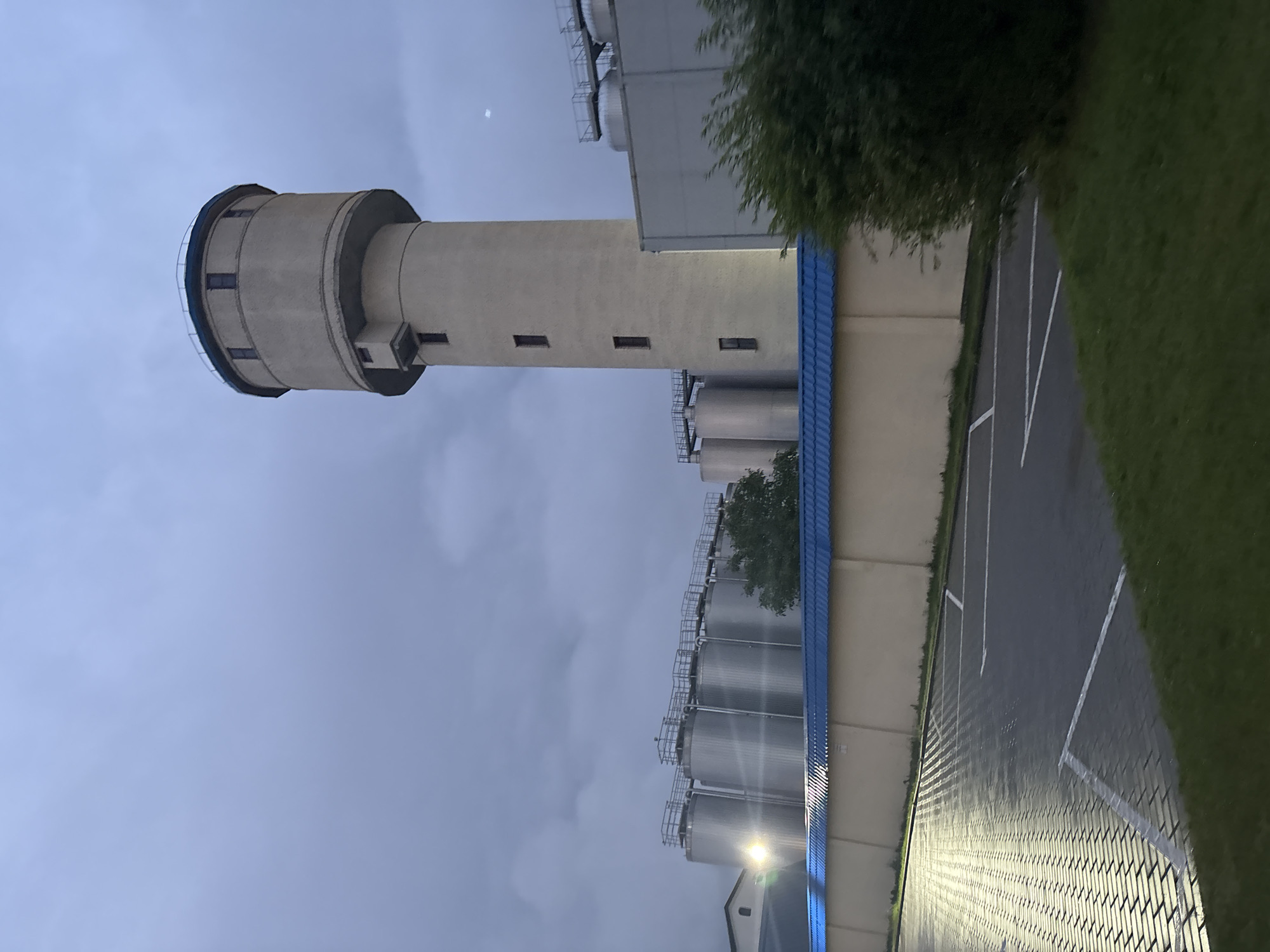This media file shows a water tower and a series of industrial silos under a cloudy sky, suggesting an overcast day or early evening. The scene appears to be an industrial complex or manufacturing facility. The water tower is a tall, cylindrical concrete structure with a rounded top and a dark band around its upper section. It has several small, rectangular windows along its height. Adjacent to the water tower are multiple large, metallic silos, arranged in rows and connected by walkways and ladders, indicating a production or storage facility. A building with a blue roof runs alongside the silos. The ground in the foreground is a paved area with white markings, resembling a parking lot, and is bordered by a strip of green grass. The lighting suggests it is either dusk or an artificially lit area, with some lights from the facility illuminating the pavement. There is no visible text in the image. The location is Lida, Belarus.