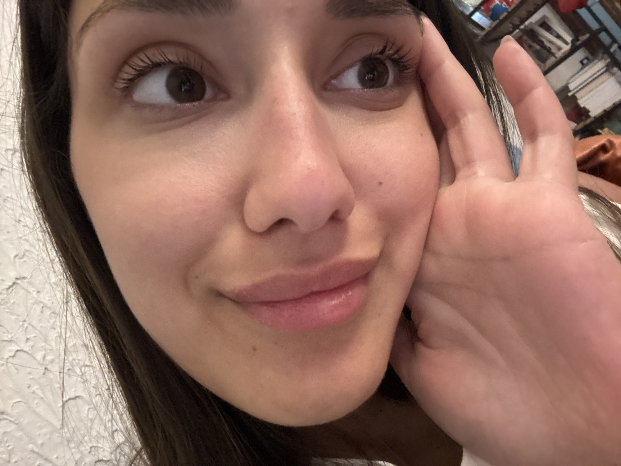 A close-up of a young woman's face, she has brown eyes, dark brown hair, and fair skin. Her hand is partially covering the right side of her face. The background is blurry, but a shelf with various objects is visible. The location in the city of Sutomore, Montenegro, is not determinable from this image alone.