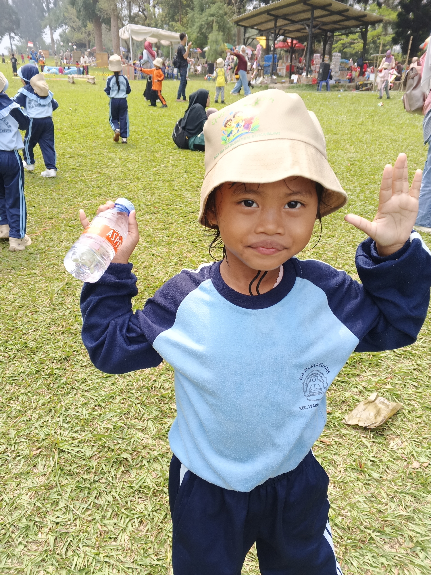 A young girl stands on a grassy field in Perbawati, Indonesia, holding a bottle of water in her right hand and raising her left hand. She is wearing a light blue long-sleeved shirt with dark blue sleeves and dark blue pants with white stripes. A tan bucket hat with an emblem of children and text partially visible, "RA NURIAELIYAH", adorns her head. The background reveals a park setting with scattered trees, a covered pavilion, and other people, some of whom appear to be children in similar uniforms. The ground is covered in short, green grass, with some dry leaves scattered about. The lighting suggests it is daytime, likely overcast or early morning due to the soft shadows.