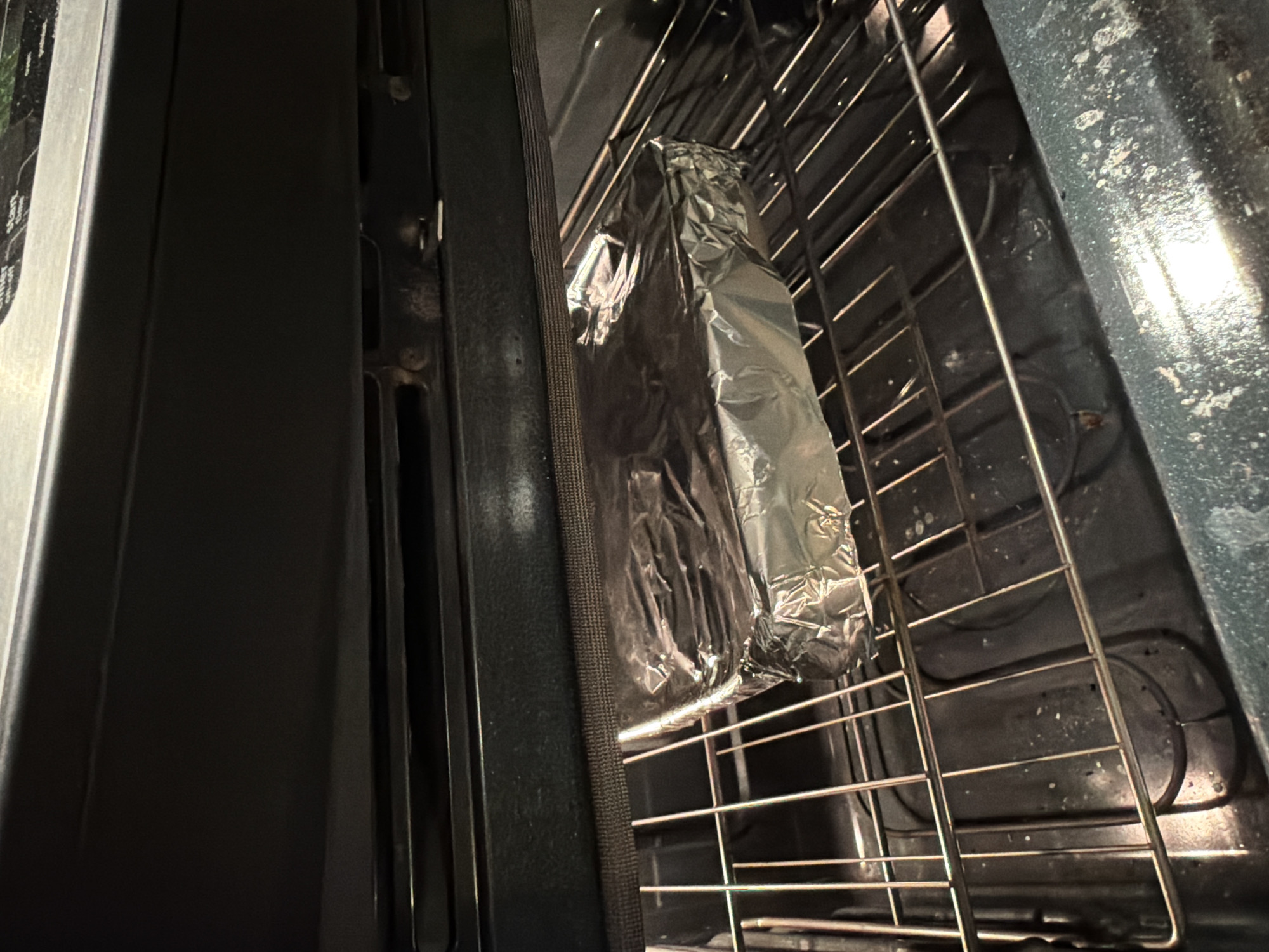 An oven interior is shown with a rack holding a rectangular package wrapped in aluminum foil. The oven door is slightly ajar, revealing a glimpse of the oven's interior. The surfaces inside the oven appear used and possibly stained. No people are visible. The location is not discernible from the image, so Montreal, Canada cannot be confirmed.