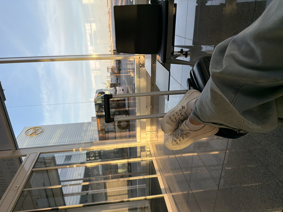 The image displays an interior view of Munich Airport (MUC), located within the Freising district of Germany. The lower half of a person is visible in the foreground, wearing light blue denim pants, white socks, and white platform sneakers with patterned laces. Their feet are elevated and resting on a black hard-shell carry-on suitcase with a silver telescopic handle. To the right, a section of black airport seating with an attached side table is visible. The terminal features large glass windows, through which a golden-hour sky, indicative of sunrise or sunset, is apparent. Outside, a multi-story glass building prominently displays a yellow Lufthansa logo. An airport bus, partially obscured but showing "Omnibusbetrieb" on its front, is parked on the tarmac, surrounded by other ground support equipment and vehicles. The polished, light-colored tiled floor of the terminal reflects the warm natural light entering through the windows.