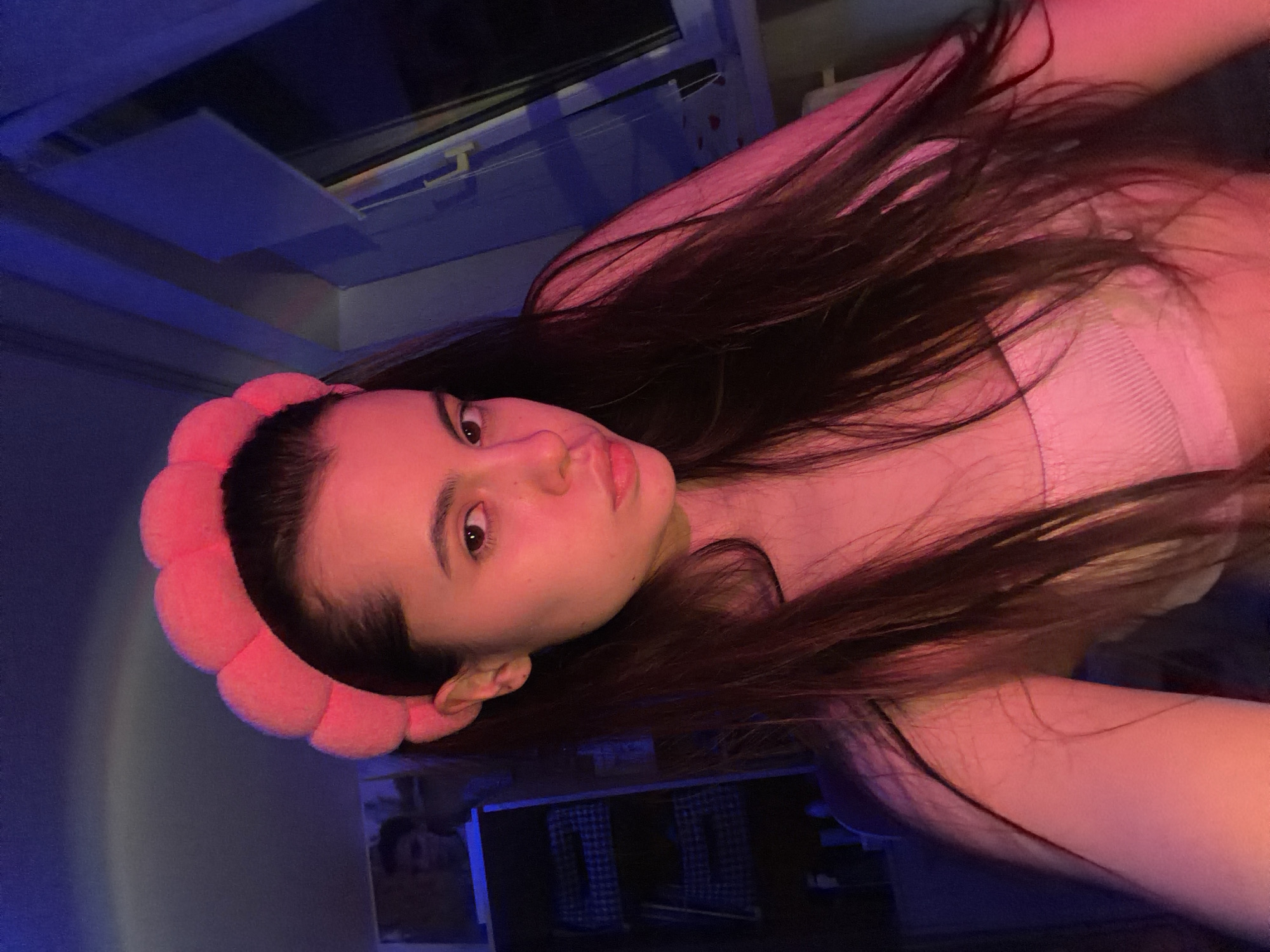 A young woman with long, dark hair is facing the camera. She is wearing a pink, flower-shaped headband. Her facial expression is neutral. She is wearing a light-colored top, possibly a tank top. The background is indistinct but appears to be a room interior with some shelving and possibly a window. The lighting is mixed, with pink and blue hues dominating the scene. There is no information in the image to confirm a location in Chaikovsky, Russia.
