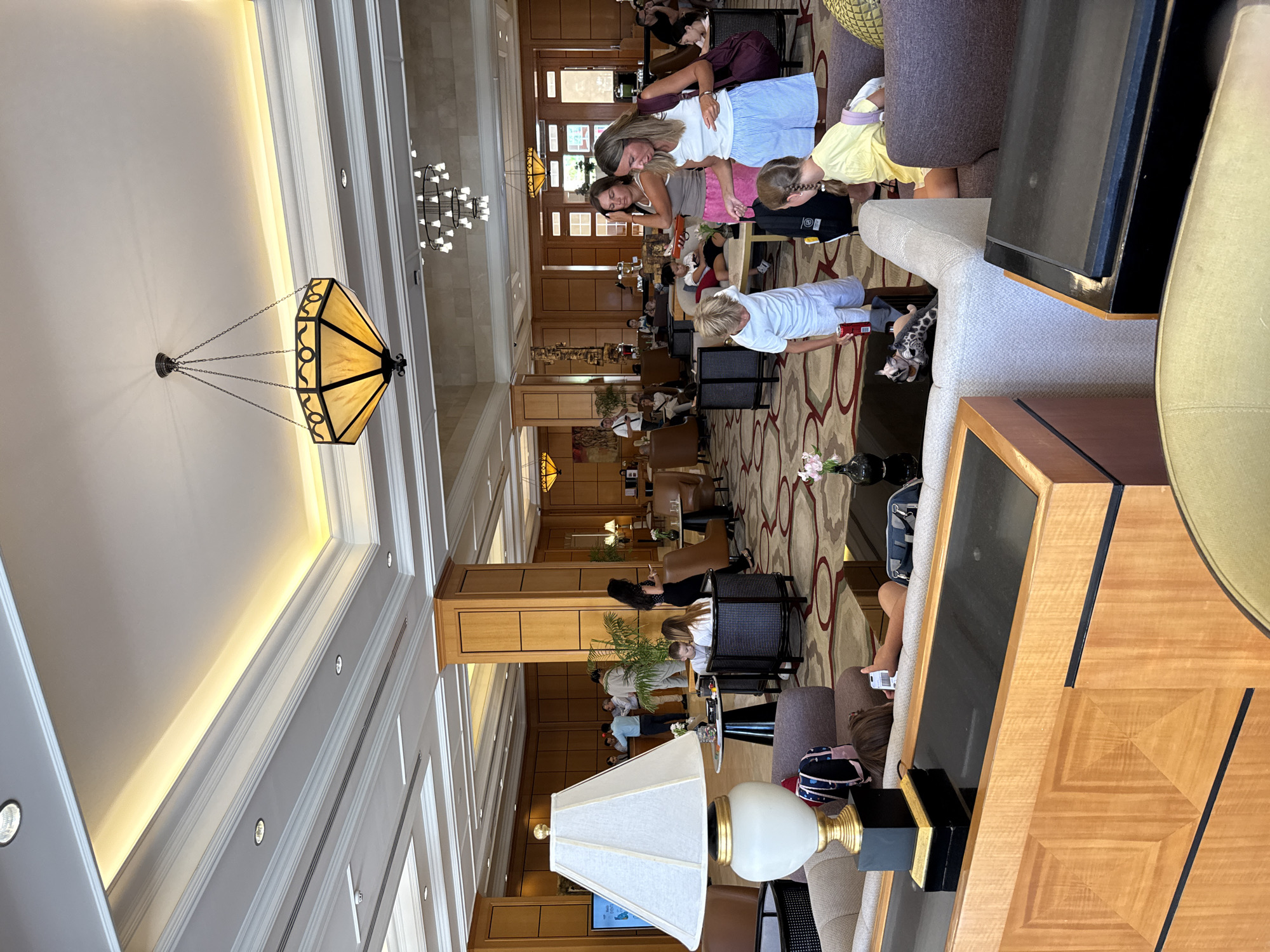 The image depicts the interior of a hotel lobby, likely in Sharm El Sheikh, Egypt, characterized by its grand architecture and comfortable seating areas. The lobby features high ceilings with decorative lighting fixtures, including a prominent hexagonal chandelier. Walls are adorned with rich wood paneling, and the floor is carpeted with an intricate, patterned rug.
Several groups of people are present, suggesting a busy hotel environment. In the foreground, a young boy in a white t-shirt and shorts stands near a sofa, holding a red can. To his right, a young girl in a yellow t-shirt sits on a sofa, her back to the camera. Two women are engaged in conversation near them, one wearing a white tank top and blue striped shorts, and the other in a grey tank top and pink shorts.
Further back, several individuals are seen mingling near the reception desk, which is visible in the background with wood paneling and screens. One person appears to be interacting with a hotel staff member. Other guests are seated in various lounge areas, some conversing and others appearing to relax.
Objects in the scene include a large table lamp with a white shade and gold accents, various upholstered sofas and armchairs in neutral tones, and small side tables. Decorative elements such as potted plants and flowers add to the ambiance. The overall impression is one of a well-appointed and active hotel lobby.