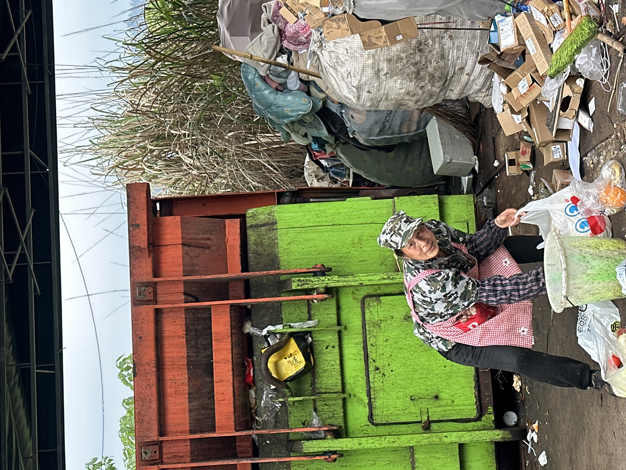 A woman wearing a camouflage hat and a red and white gingham apron is working beside a large, orange and green dumpster. The dumpster is filled with various items, including cardboard boxes, plastic bags, and textiles. The area around the dumpster appears littered, with scattered debris. Tall, dry grass grows nearby. The location is Jiangjin, China.