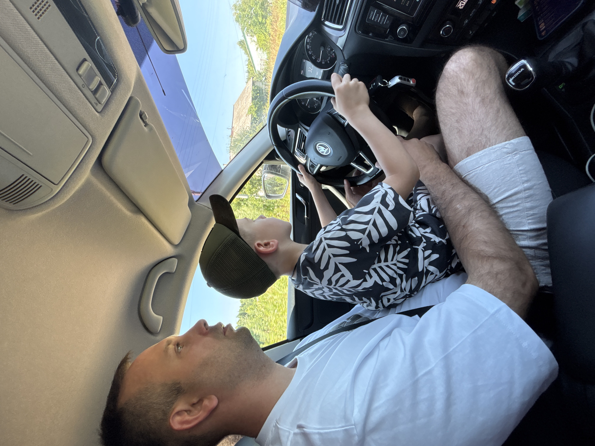An adult male, wearing a white t-shirt and white shorts, is seated in the driver's seat of a car. He is looking to the left, with his right arm resting on his lap and his left arm partially visible. A young boy, wearing a dark t-shirt with a white leaf pattern and a dark baseball cap, is seated in front of him and is holding the steering wheel, appearing to drive the car. The boy is looking forward. The car interior shows a dashboard with a speedometer indicating 32, a radio, and other controls. Outside the car, through the windshield and side windows, a rural landscape is visible, with houses and trees under a clear sky. The location is identified as Dunilovo, Russia.
