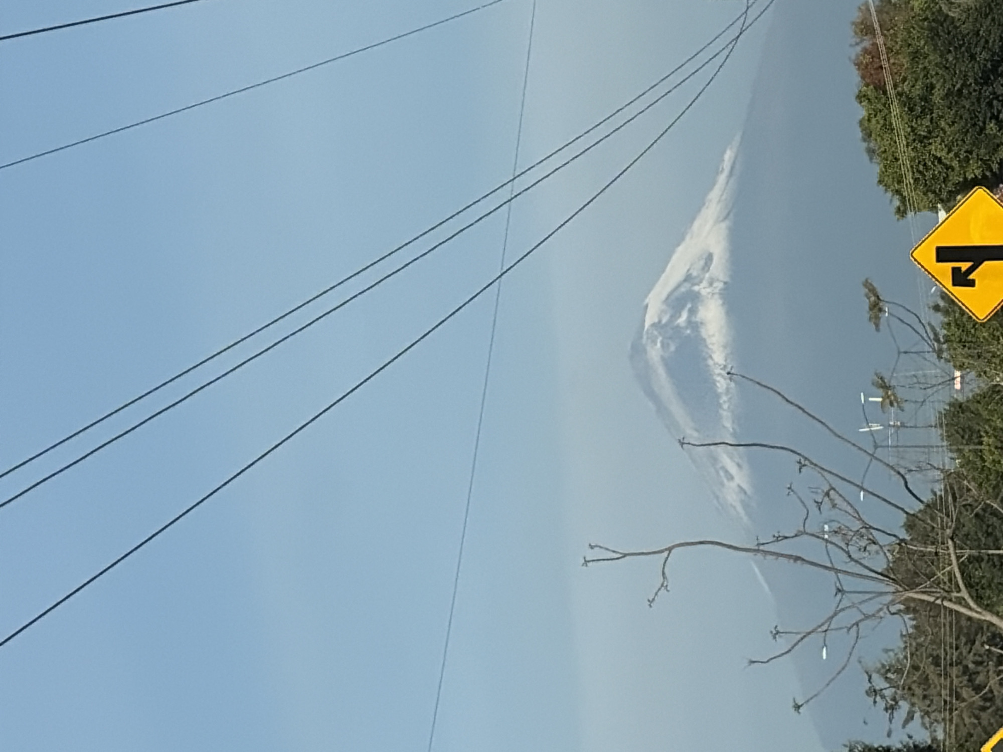 The image shows a view in Puebla, Mexico, likely near a road. In the background, there is a mountain partially obscured by haze or clouds. In the foreground, there are power lines, tree branches with few leaves, and a yellow road sign indicating a junction. Green trees are visible on the right side of the frame. The sky is a clear blue.