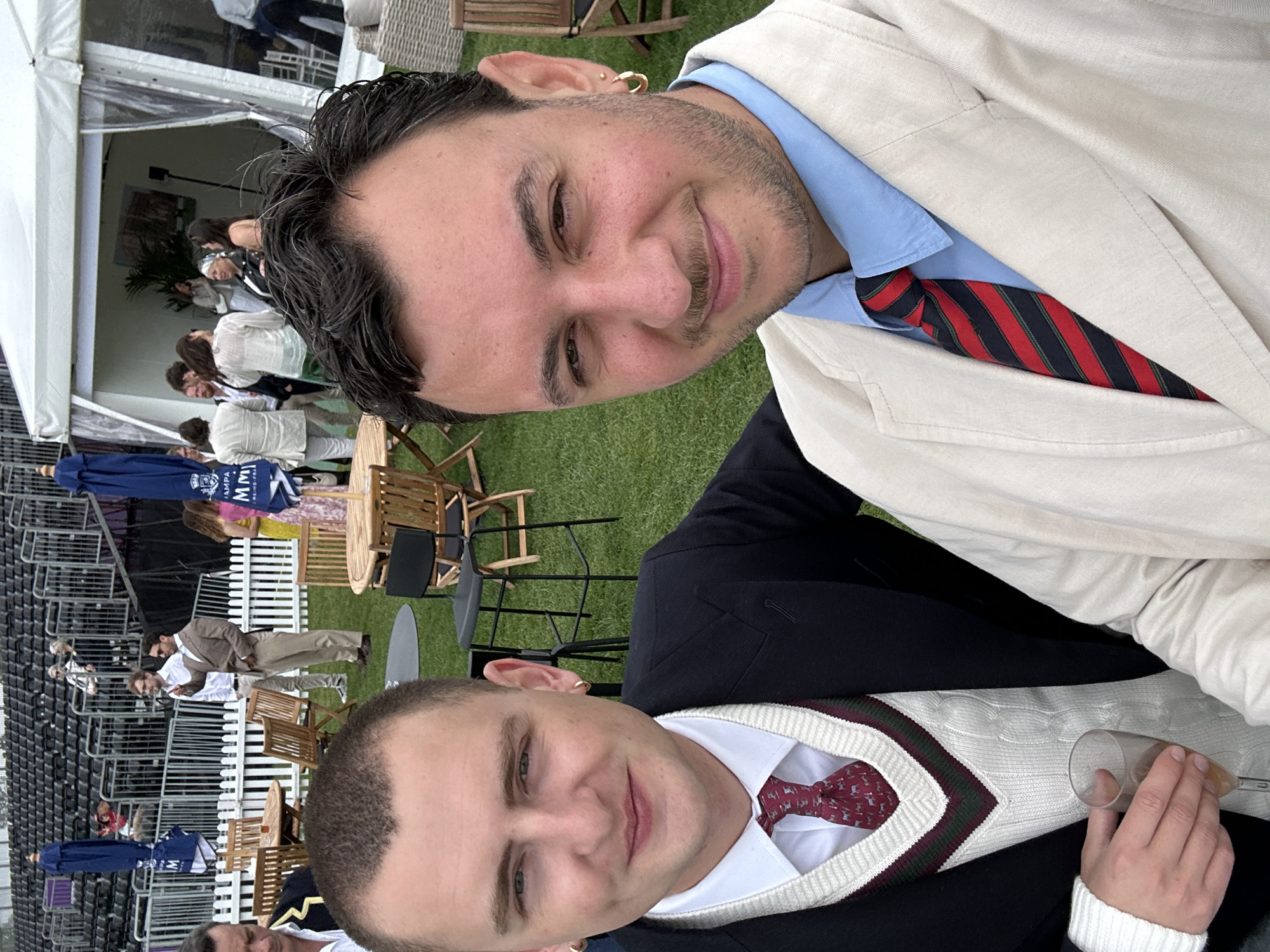 The image depicts an outdoor hospitality area at an event in the city of London, United Kingdom. Two men are captured in a close-up selfie in the foreground, smiling faintly and looking directly at the camera.
The man on the left has short, light brown hair, light eyes, and wears a dark navy blazer over a white collared shirt. He sports a white cable-knit V-neck sweater vest with horizontal maroon and dark green stripes on the V-neckline. His tie is maroon with a repeated pattern, possibly small animals. A single gold hoop earring is visible in his left ear. He holds a stemmed glass containing a light amber liquid, likely champagne, in his right hand.
The man on the right has dark, curly hair and a trimmed beard/stubble. He has light eyes and wears a light beige blazer over a light blue collared shirt. His tie is striped in red, black, and thin green. A single gold hoop earring is visible in his right ear.
The background shows a lively outdoor event setting. Green grass covers the ground, furnished with numerous round wooden tables and matching chairs, as well as some black high-top tables with stools. A white picket fence runs across the midground, separating the immediate area from further elements. Beyond the fence, a large tiered stadium or grandstand with dark seating is visible, occupied by spectators. Blue market umbrellas, some bearing "CHAMPAGNE MM" branding, are open above seating areas. To the right, a large white marquee tent with clear plastic side panels is present, with several individuals mingling inside and outside. One section inside the tent shows a potted plant and a framed picture on the wall. The overall ambiance suggests a social gathering at a sporting or entertainment event.