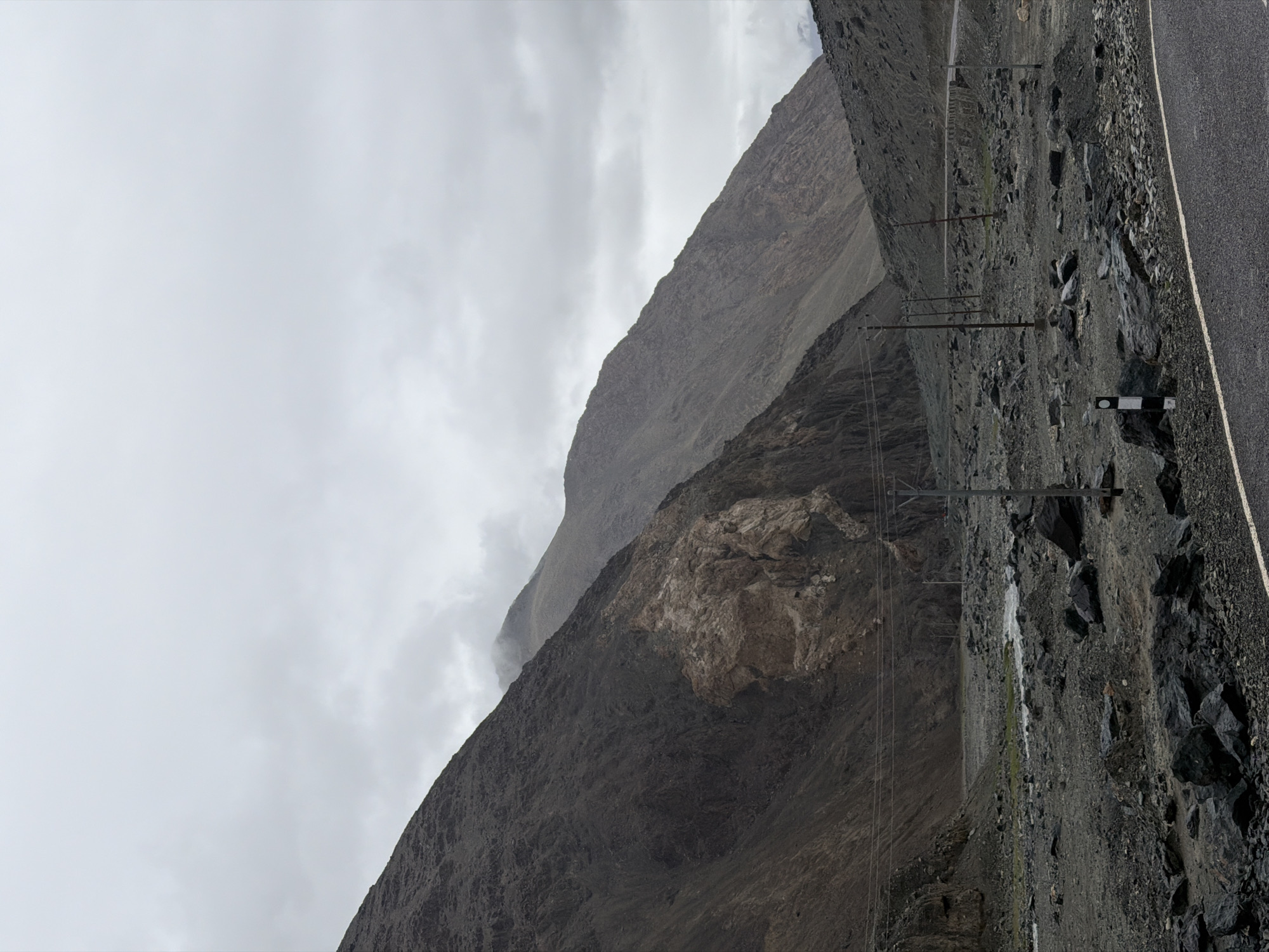 The image depicts a rugged, mountainous landscape with a cloudy sky overhead. The terrain is characterized by steep, rocky slopes and a barren, stony valley floor. A paved road curves through the scene, with a white line marking its edge. Several power poles with wires are visible, indicating human infrastructure in this remote area. In the distance, a small stream or river flows through the valley. The overall atmosphere is somber and desolate. The location is identified as Sumdo, India, a village in the Ladakh region known for its stark, high-altitude environment. No people are present in the image.