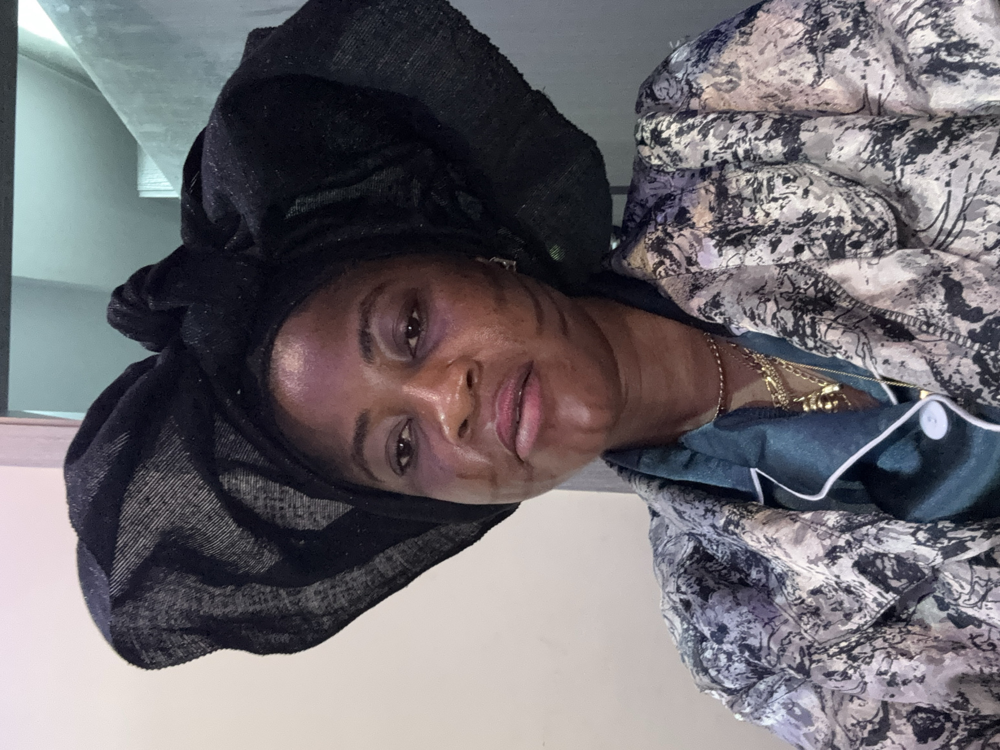 This is a portrait of a woman wearing a black headwrap and a patterned robe, indoors. The woman is looking towards the camera with a slight smile. Her face shows some makeup, with visible eyeliner and lipstick. She is also wearing a gold necklace. The setting appears to be a domestic environment, possibly a bedroom or living area, with a neutral-colored wall in the background and what looks like a shelf or ceiling above her. The lighting suggests it could be daytime, but there are no strong indicators of the specific time. The overall mood is casual and relaxed. No text is visible in the image.
