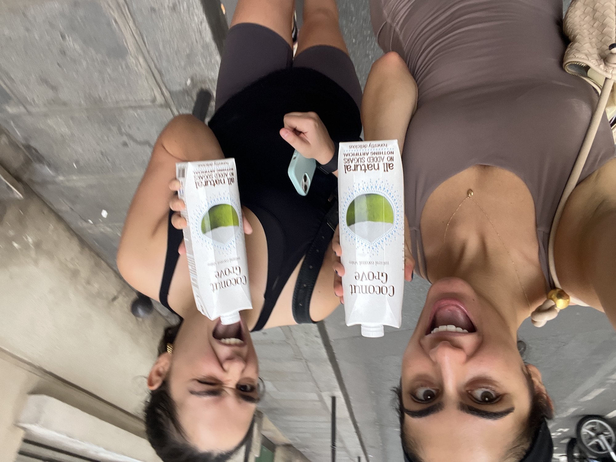 Two young women are holding cartons of Coconut Grove natural coconut water. The woman on the left, wearing a taupe tank top and a gold necklace, is looking at the camera with her mouth open and eyes wide. The woman on the right, wearing a black tank top and grey shorts, is winking and has her mouth open. She is holding a light blue phone in her right hand. Both women are on a street in Paris, France, with buildings visible in the background and a cobblestone street beneath them. A motorcycle is parked to the left of the frame. The lighting suggests it is daytime.