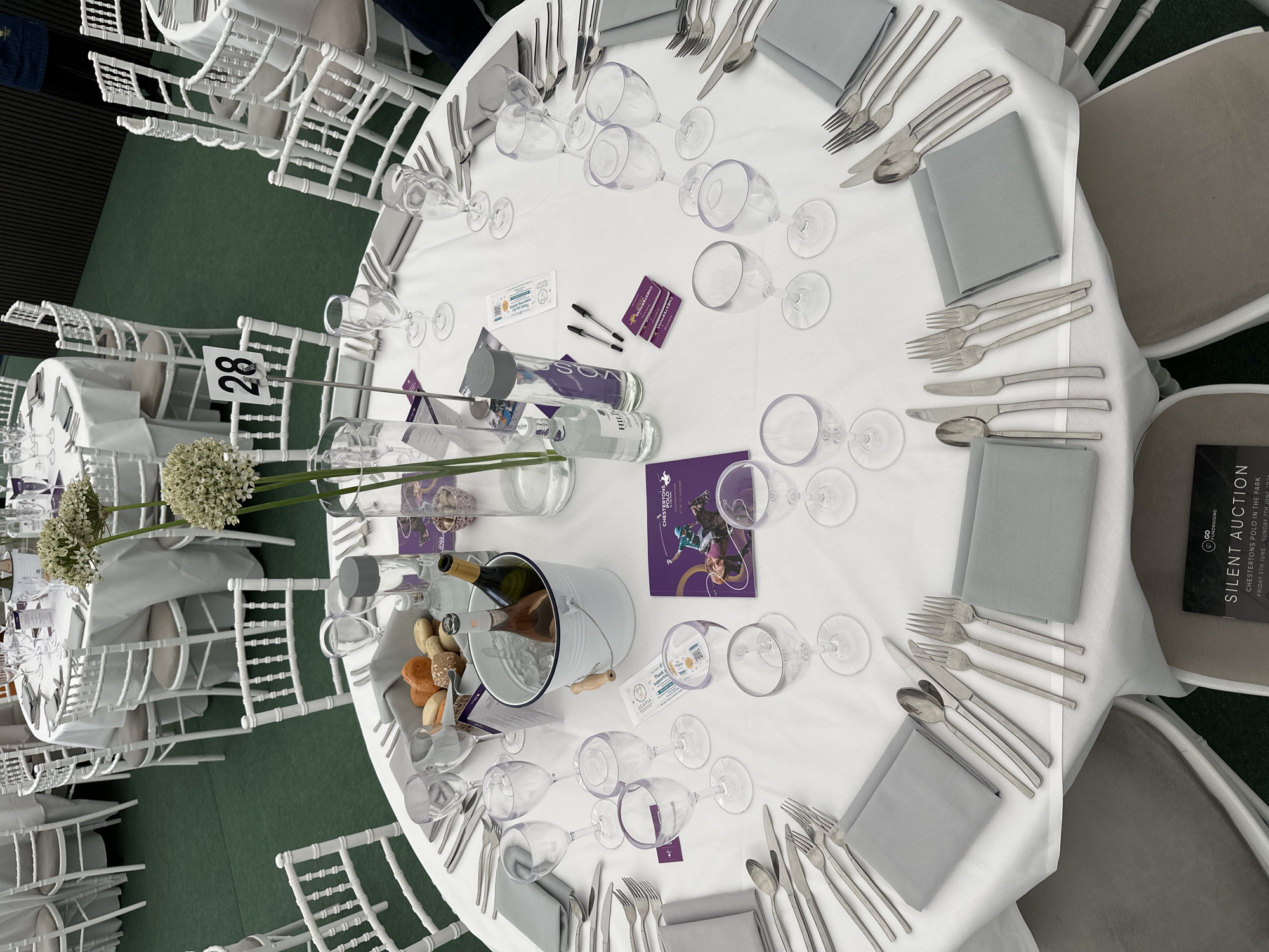 This image depicts a formal event setting at Chestertons Polo in the Park, located at Hurlingham Park in Fulham, London, United Kingdom. The scene shows an event space featuring a green carpeted floor, mimicking grass. Multiple round tables, draped in white tablecloths, are arranged throughout the area, surrounded by white Chiavari chairs with light beige cushions.
The foreground focuses on one such round table set for multiple guests. Each place setting includes a light gray fabric napkin, a complete set of silverware (spoon, knife, and two forks), and an assortment of clear plastic wine and water glasses. Centered on the table is a white metal ice bucket containing two bottles of wine—one rosé and one dark-colored bottle. Adjacent to the bucket, a tall clear vase holds two stems of spherical white flowers. A bread basket filled with various bread rolls is also present. Other items on the table include a white "28" table number sign on a metal stand, two water bottles (one VOSS and one "HILL" branded), two black pens, and various purple-themed event materials. These materials include brochures titled "CHESTERTOWNS POLO IN THE PARK" (one displaying a polo player and dated "FRIDAY 5TH JUNE - SUNDAY 7TH JUNE 2020"), "SILENT AUCTION" literature, and small branded cards.
In the background, other identically set tables with chairs are visible. A person wearing a dark blue garment is partially visible standing behind a table at the far right edge of the frame. No other individuals are seated or actively engaging with the table settings, indicating the scene is set prior to guest arrival.