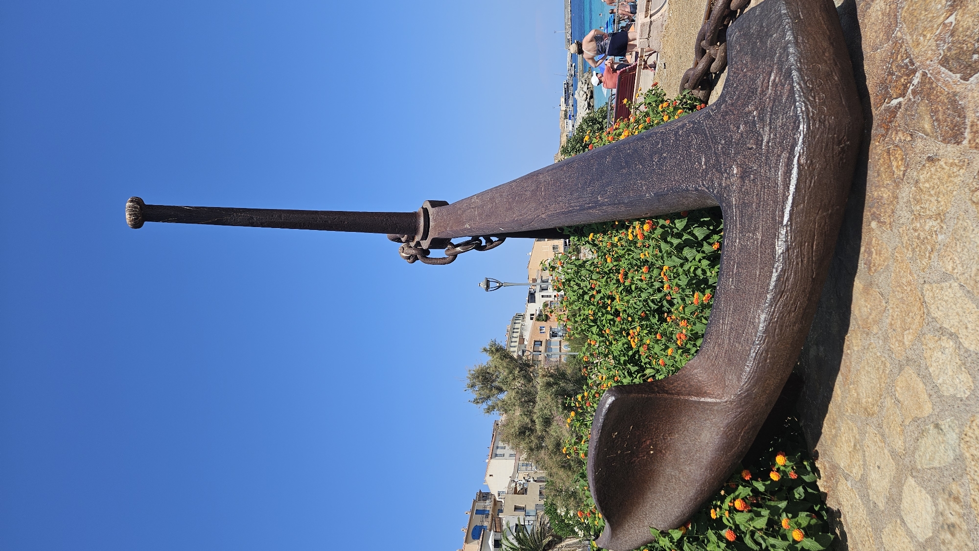 The image captures a large, weathered, brown metal anchor displayed in L'Île-Rousse, France. The anchor is positioned in the foreground, with its fluke resting on a stone pathway and its shank extending upwards against a clear blue sky. Behind the anchor, a vibrant bed of orange and yellow flowers borders the scene. In the middle ground, a line of buildings with light-colored facades and terracotta roofs forms the town's backdrop, with some trees and a lamppost interspersed. The far background reveals a glimpse of the sea and a few boats, suggesting a coastal setting. A few people are visible in the distance, seated on a bench or walking along the beach. The scene is illuminated by bright sunlight, indicating it is daytime.