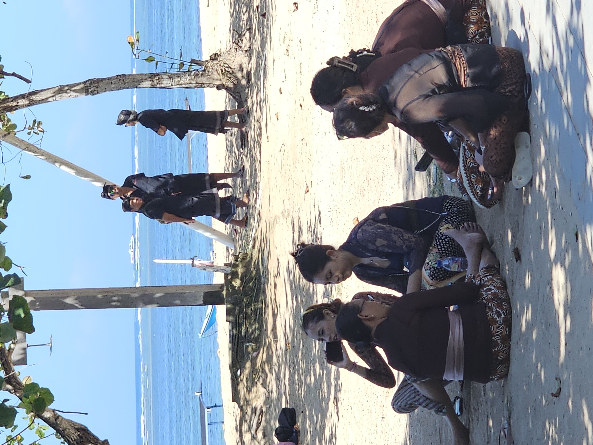 The image depicts a group of people on a sandy beach, likely in Denpasar, Indonesia, with the ocean and a clear blue sky in the background. Several individuals are seated on the sand, some of whom are wearing traditional Balinese attire, including batik sarongs and dark, patterned tops. One woman in the foreground is holding a phone to her ear. Further back, a group of men in black clothing and headbands stand under a tree, overlooking the water. The environment suggests a sunny day, with sunlight filtering through tree branches, casting shadows on the sand. The sea is visible with light waves and a distant boat.
