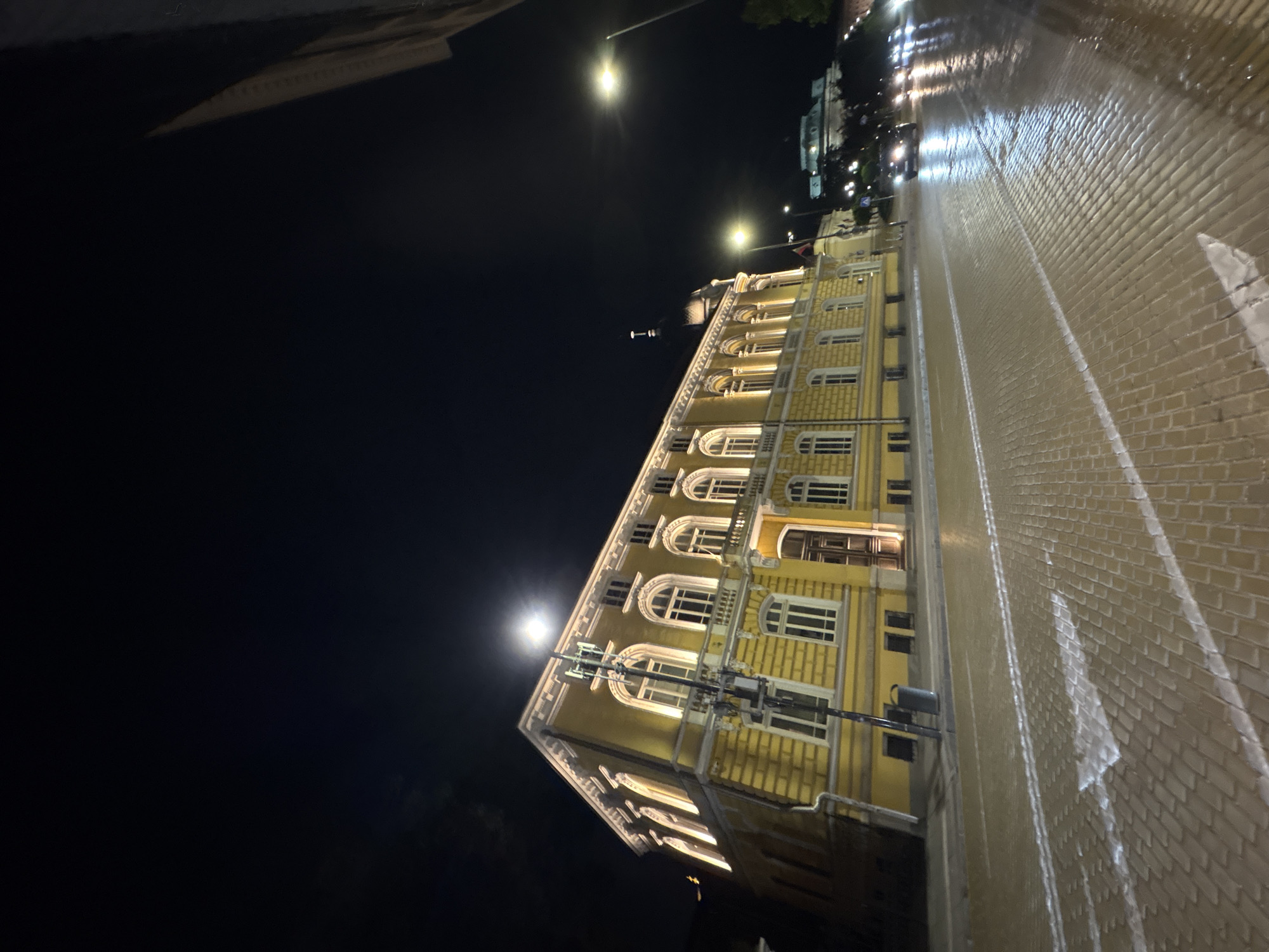The image captures a nighttime urban scene, with a prominent building on the left and a wet street extending into the distance on the right. The time of day is clearly night, indicated by the dark sky and extensive artificial lighting.
The main subject is a multi-story building with a vibrant yellow facade, characteristic of classical European architecture. Its exterior is brightly illuminated by numerous lights, highlighting its architectural details such as ornate cornices, decorative window frames, and a series of arched windows on the upper floors. Other windows are rectangular. Wires or cables are visible running along a portion of the building's exterior.
To the right of the building, a paved street, likely made of bricks or cobblestones, stretches away. The street appears wet and highly reflective, suggesting recent rain. It reflects the overhead streetlights and the building's illumination, creating streaks of light. Faint white road markings, including what appears to be an arrow shape, are visible on the wet surface.
The sky above is dark black, contrasting sharply with the bright artificial lights. Several streetlights emit strong glows, and a distinct, bright, and circular light source, resembling a full moon, is visible in the upper left portion of the sky, casting a soft glow. In the far distance, beyond the main building and street, another structure with some lights can be faintly seen, possibly another building or monument.
The scene is devoid of clearly identifiable people or vehicles, contributing to a sense of quietude and stillness. The wet street implies recent weather activity. No visible text can be discerned in the image.