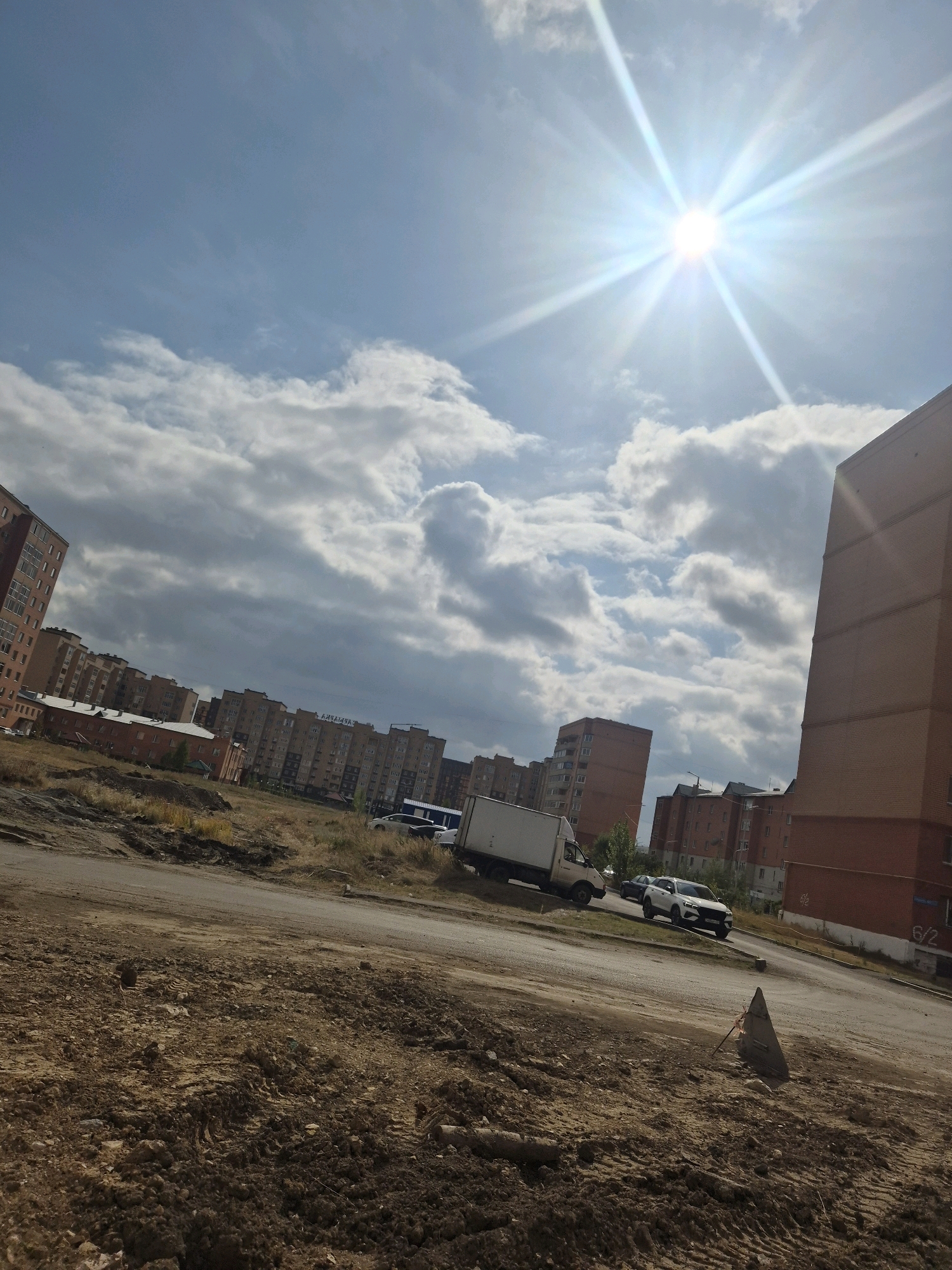 The image depicts an urban scene in Kokshetau, Kazakhstan, on a sunny day with scattered clouds. In the foreground, there is a patch of dry, uncultivated land with loose soil, indicative of construction or a similar disturbance. A dirt road or pathway runs through this area.
In the middle ground, a white truck is parked on the right side of the road, facing left. To its right, a white SUV is driving away from the viewer. Further down the road, more vehicles and residential buildings are visible.
The background is dominated by a series of multi-story apartment buildings, primarily constructed of brick. These buildings extend across the horizon, suggesting a residential district. The sky is bright blue with several large, fluffy white clouds. The sun is shining intensely, creating a lens flare effect in the upper right quadrant of the image.
The overall impression is one of a typical day in a developing or redeveloping urban area, with construction in progress alongside established residential structures. No people are clearly visible in the image.