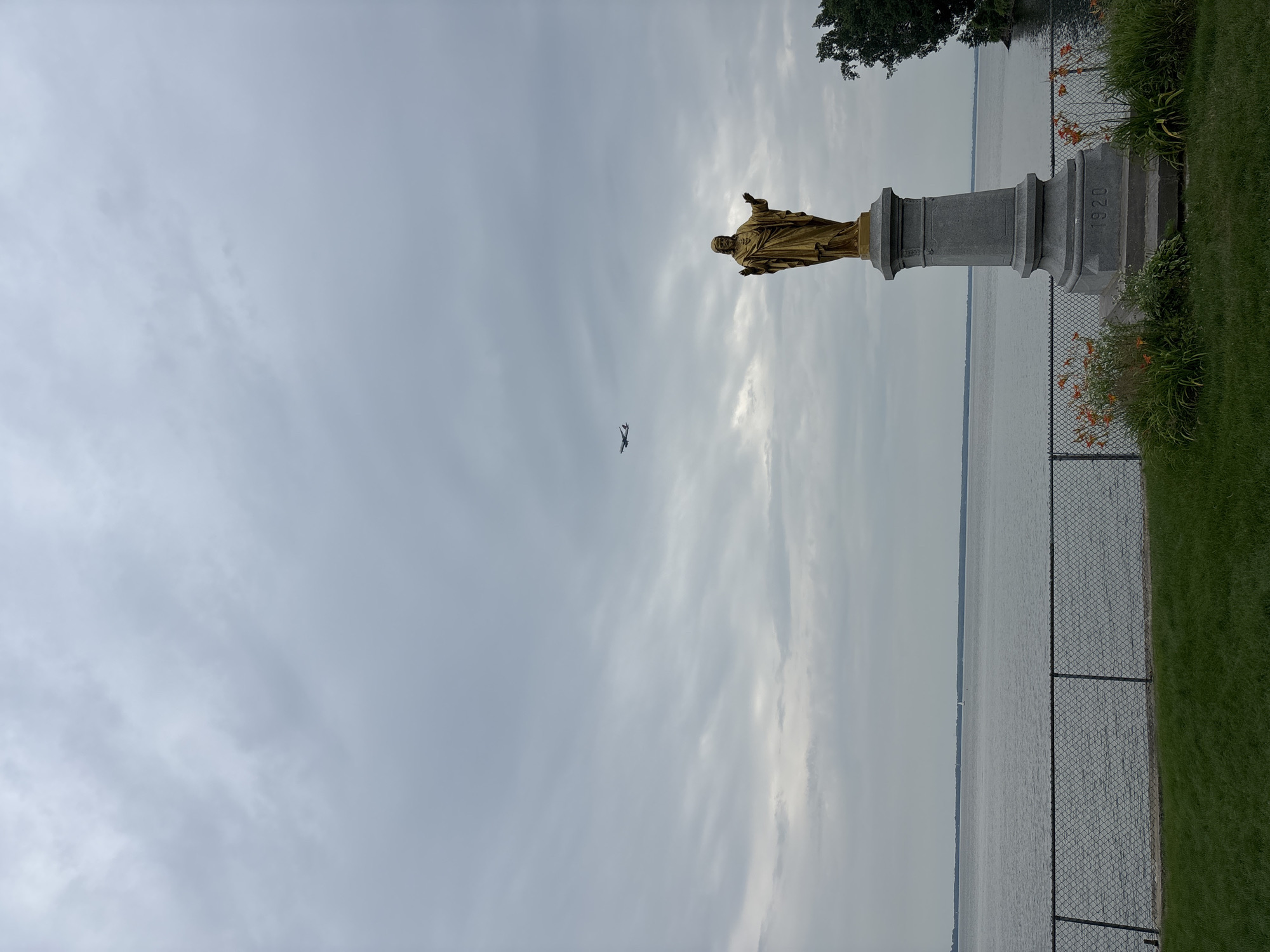 The image depicts a golden statue of Jesus Christ standing on a grey stone pedestal, situated in front of a large body of water. The statue faces forward with arms outstretched, and the pedestal is inscribed with the year "1920." A chain-link fence runs horizontally across the foreground, separating a manicured green lawn from a grassy area adorned with orange flowers and greenery. In the distance, an airplane is visible flying in the cloudy sky. The water appears calm, with a sailboat in the far distance. The overall environment suggests a park or memorial site, possibly overlooking a lake or river. This statue is located at the Pointe-Claire Yacht Club in Pointe-Claire, Canada.