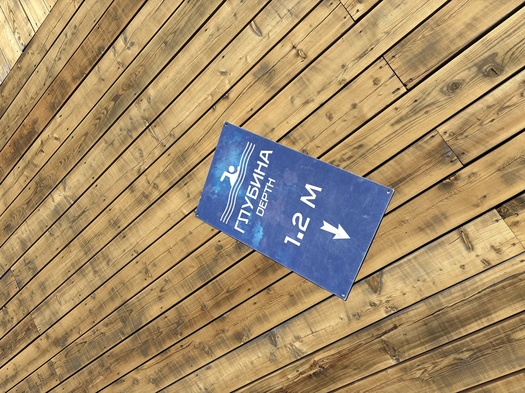 An overhead, slightly angled photograph showcases a blue rectangular sign placed on a wooden deck. The deck consists of numerous light brown wooden planks, arranged diagonally across the frame. Bright sunlight illuminates the scene, casting distinct shadows between the planks and indicating a clear, sunny day, likely midday or early afternoon. The wooden surface displays natural grain patterns, visible knots, and some areas with darker weathering, with exposed nails or screws securing the planks.
The blue sign features white text and an icon providing information about water depth. On the left side, a stylized icon of a swimmer is depicted amidst wavy lines, symbolizing water. Vertically written in Cyrillic script below the icon is "ГЛУБИНА" (GLUBINA), which translates to "DEPTH", followed by the English word "DEPTH" written horizontally. To the right, the numerical measurement "1.2 M" (1.2 Meters) is displayed, accompanied by a prominent downward-pointing white arrow.
No people are visible in the image. The presence of the depth sign strongly suggests proximity to a swimming pool, lake, or other body of water where depth information is relevant for safety or recreational purposes. The Cyrillic text on the sign indicates a location in a Russian-speaking region, consistent with the provided context of Дрокино, Russia. The image is a factual depiction of an informational sign on a wooden surface.