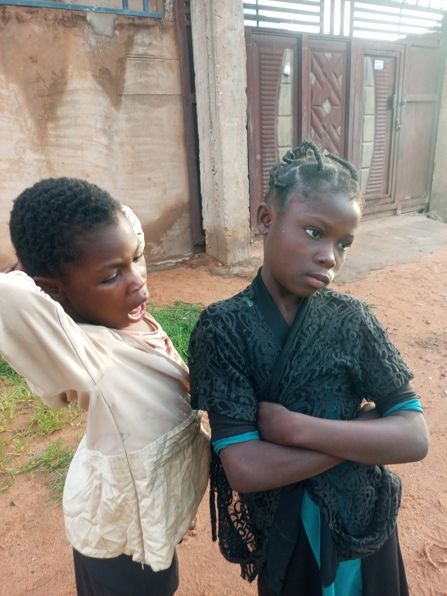 Two young girls are standing outdoors in what appears to be a dusty, unpaved area, possibly in Tempure, Nigeria. The girl on the left is seen from the chest up, facing towards the right. She has dark, short hair and is wearing a light-colored jacket with a zipper and a pink undershirt. Her mouth is open, and her left arm is raised behind her head, with her hand touching the back of her head. The girl on the right is facing towards the left, with her arms crossed. She has braided dark hair and is wearing a dark, possibly black, lacy top over a teal garment, and a black skirt. The background consists of a rough, textured wall on the left, and a large, dark wooden gate with horizontal slats and decorative panels behind the girls. The ground is sandy and reddish-brown, with patches of dry grass. The lighting suggests it is daytime.