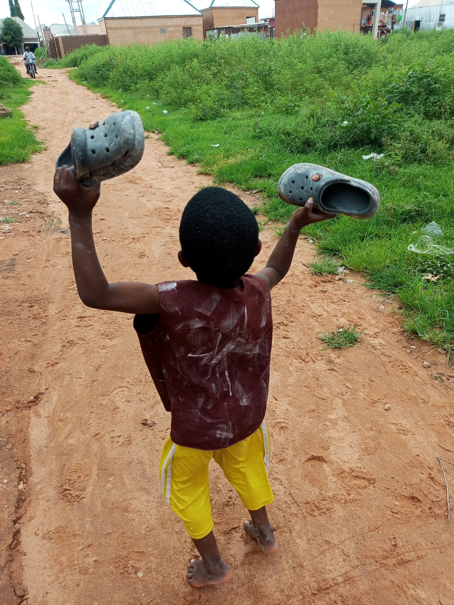 In a dusty, unpaved lane in Tempure, Nigeria, a young boy, viewed from behind, raises a gray Croc shoe in his left hand and another in his right, each held aloft with outstretched arms. He wears a dark maroon t-shirt, visibly stained with white paint splatters, and bright yellow shorts with white stripes. His dark hair is short and neatly cut. The lane is flanked by dry, sandy earth and patches of vibrant green grass. In the background, modest buildings with earth-toned walls and corrugated metal roofs are visible, interspersed with dense green foliage. The sky is overcast, suggesting a cloudy day. Further down the lane, a man on a motorcycle rides away from the viewer. The overall scene depicts a rural or semi-rural setting within Tempure.