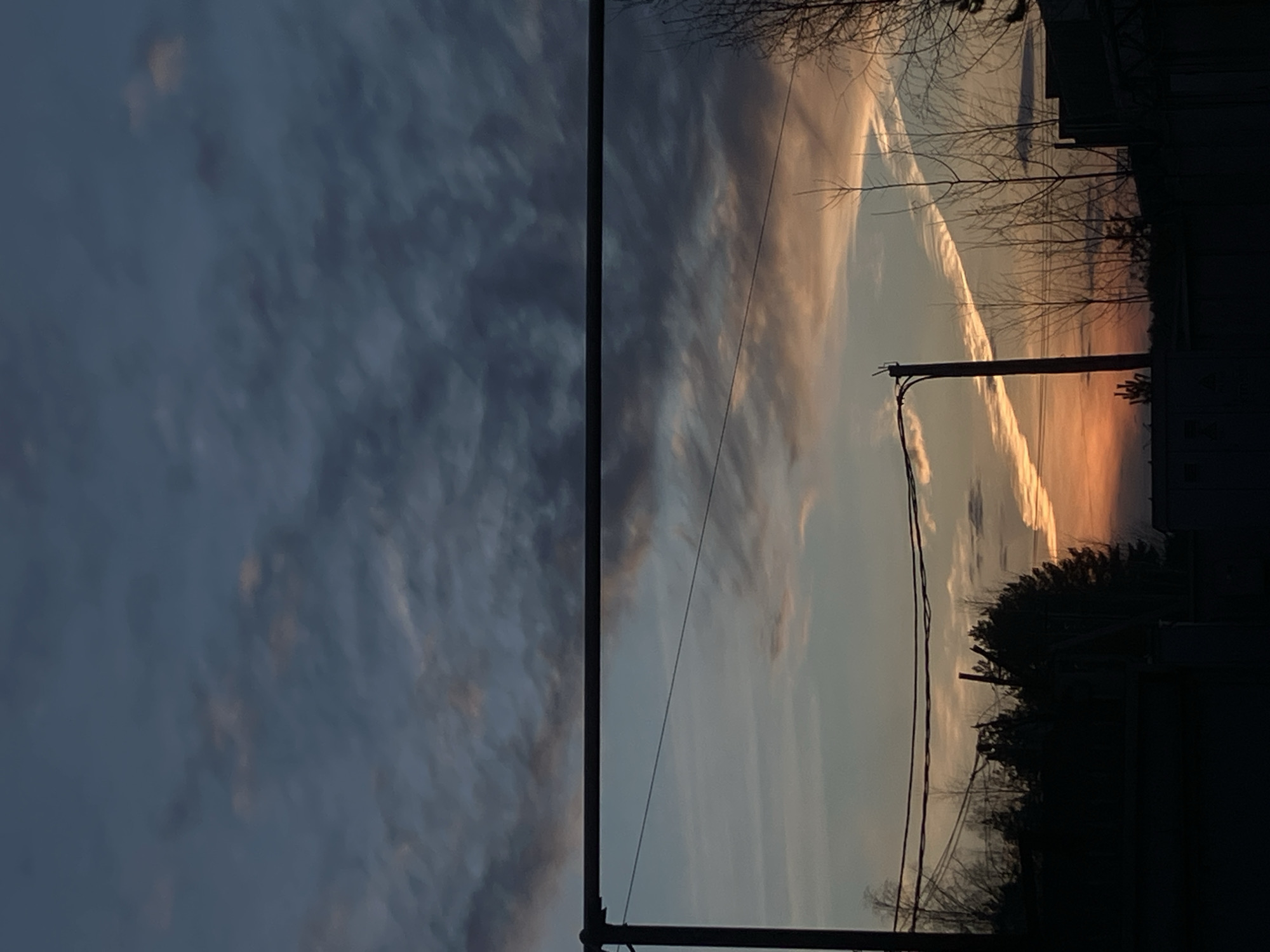 The image captures a view of the sky, bisected vertically by a utility pole. The left side of the frame shows a predominantly gray, overcast sky with scattered cloud formations. The right side of the image reveals a sky illuminated by a setting sun, with hues of orange and yellow visible through the clouds. Bare tree branches are silhouetted against the sunset sky. In the lower right corner, the dark outlines of buildings and trees are visible, suggesting a residential area. Power lines run across the frame, adding to the urban setting. The photograph appears to be taken at dusk or dawn. No people are visible in the image. Given the orientation, the location in the city of Noviy, Russia, is not clear.