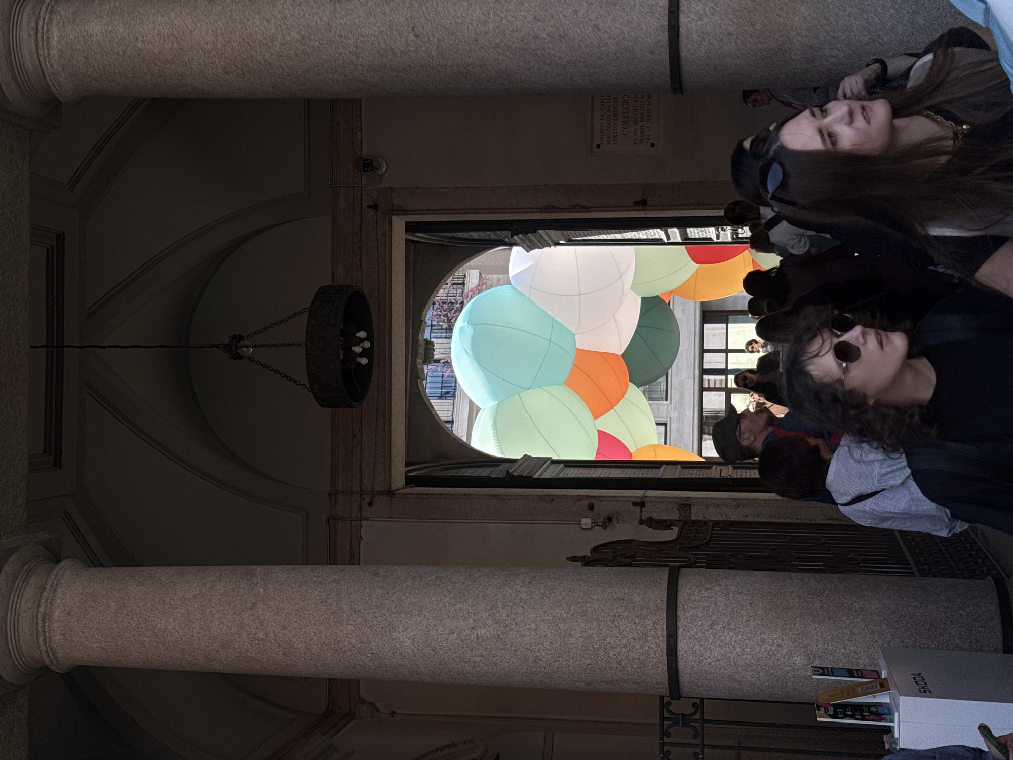 The image depicts the interior of a historic Italian building, likely a college or cloister, with people observing a colorful art installation. The perspective is slightly angled, looking down into a space with classical architectural elements.
In the foreground on the right, several individuals are gathered. A young woman with dark curly hair and sunglasses looks towards the center, while behind her, another woman with long dark hair gazes upwards and to the left, seemingly engaged by the scene. Other figures are visible in the background, including a man wearing a cap.
The setting is characterized by stone columns, including a prominent one in the lower half of the frame, and arched ceilings. A dark, ornate chandelier hangs in the upper left. Through an archway in the center, a vibrant art installation is visible, composed of numerous large, colorful inflatable spheres in shades of light blue, white, orange, green, and pink. These balloons create a striking modern contrast against the traditional architecture and bright light pouring in from beyond the archway.
On a white box in the lower right, the brand name "SKODA" is visible, alongside some books or magazines. A stone plaque mounted on the wall above the archway bears Italian text, including "VOLUTO DA CARLO," "REALIZZATO DA FEDERICO," "QUESTO EDIFICIO," "COLLEGIO," "FU NEI SECOLI XVI," and "TRA LE TERRE LOMBARDE," indicating that the building is a historical college from the 16th century in the Lombard region. The scene captures a moment of public engagement with contemporary art within a significant historical context in Milan, Italy.